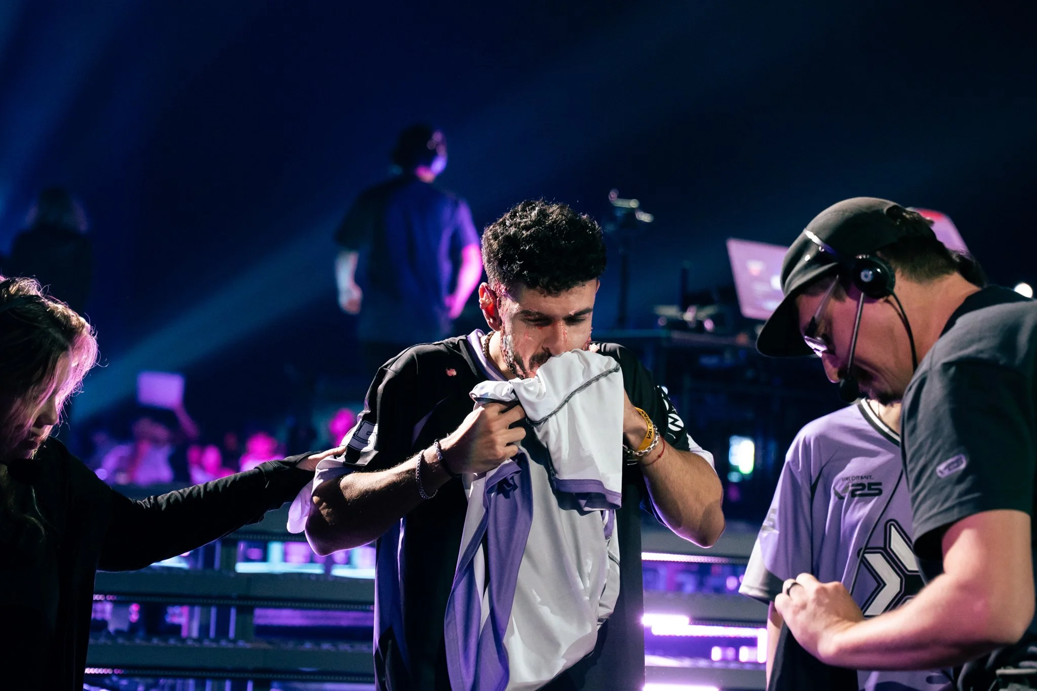 Three people, a man with short dark hair and a beard, and two with long and short hair, are on a stage with purple and blue lighting. The man is wiping his face with a white cloth, seeming emotional, while others tend to him. There is a man with glas