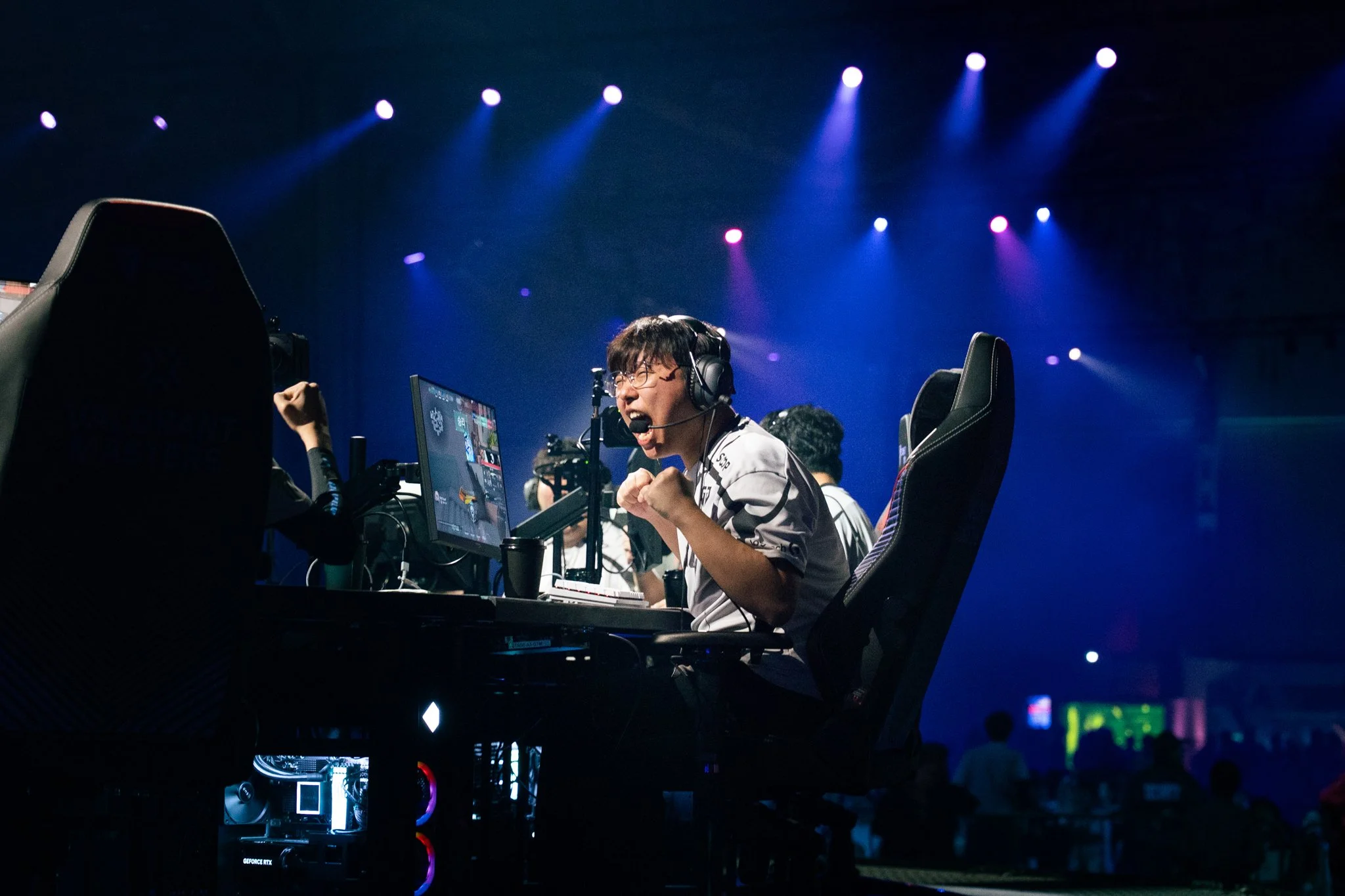 E-sports player celebrating at a gaming tournament with illuminated stage lighting and gaming equipment.