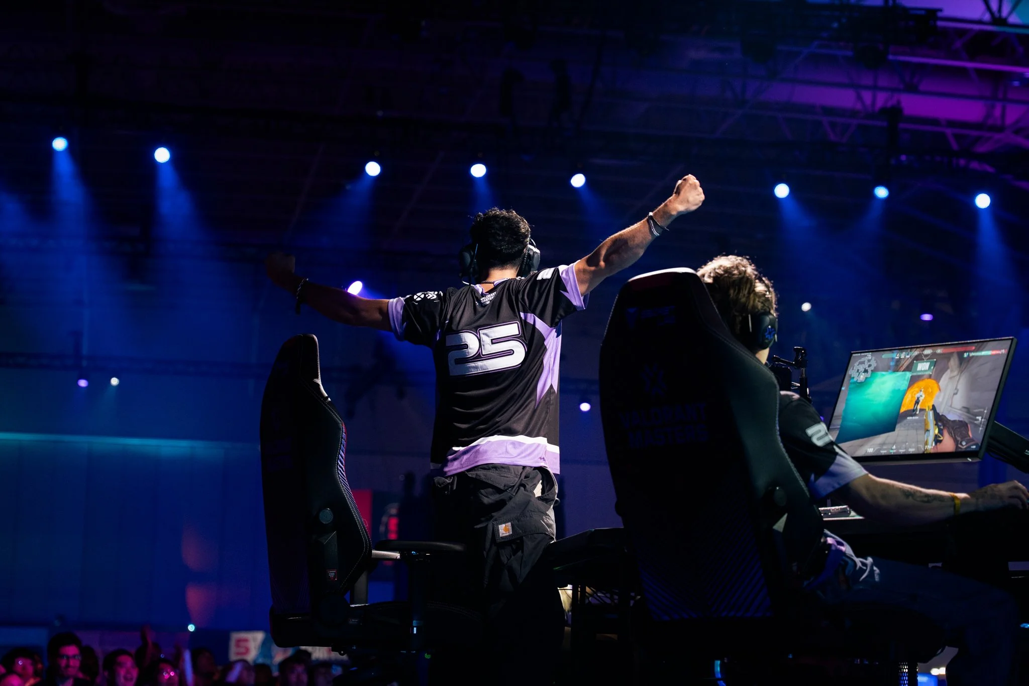 An esports player wearing a black jersey with purple accents and the number 25 celebrates with arms outstretched after a win during a gaming tournament, with another player at a computer monitor displaying gameplay.