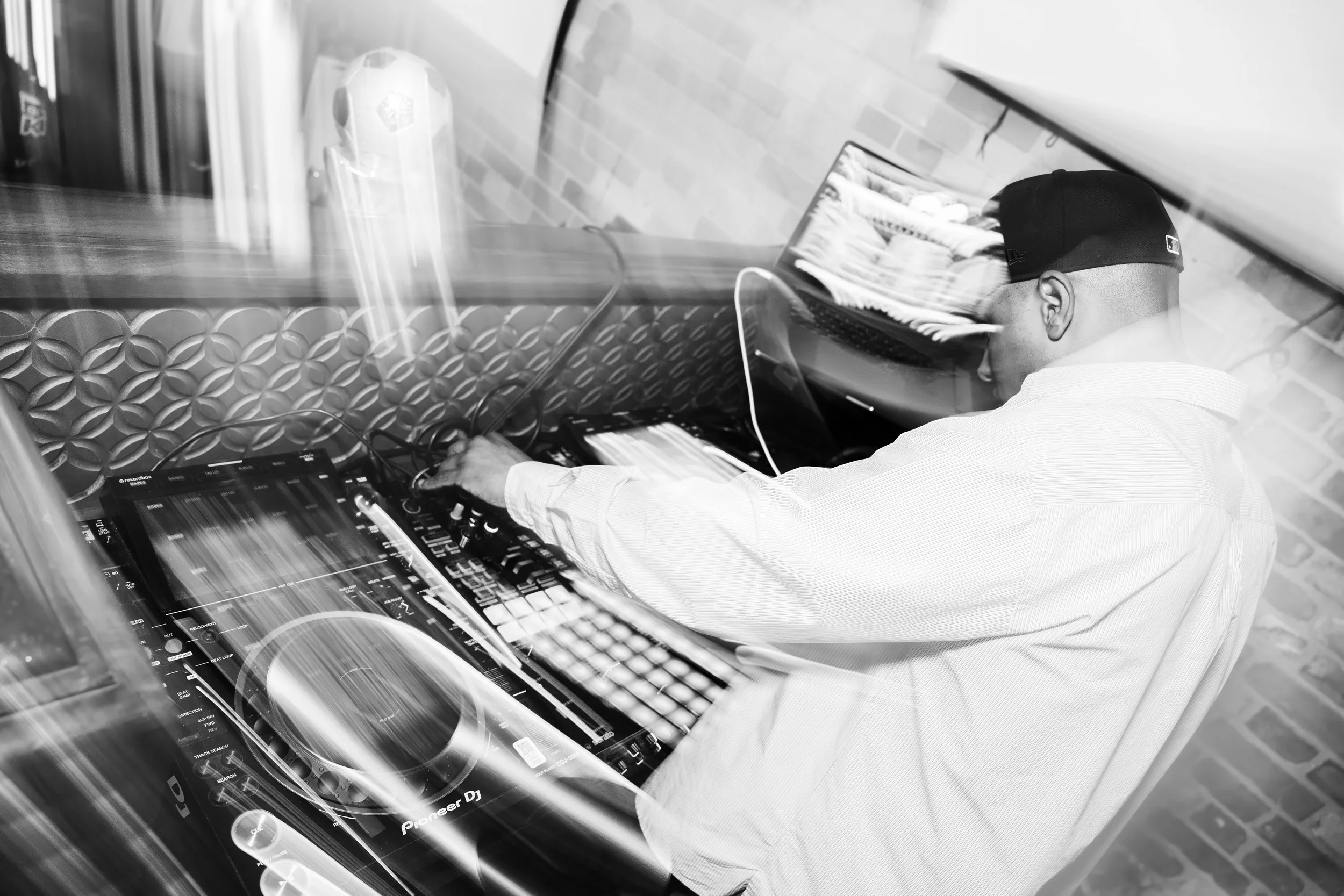 A DJ wearing a baseball cap and a button-up shirt, working with a DJ controller and a laptop, in a setting with brick and patterned wall decor.