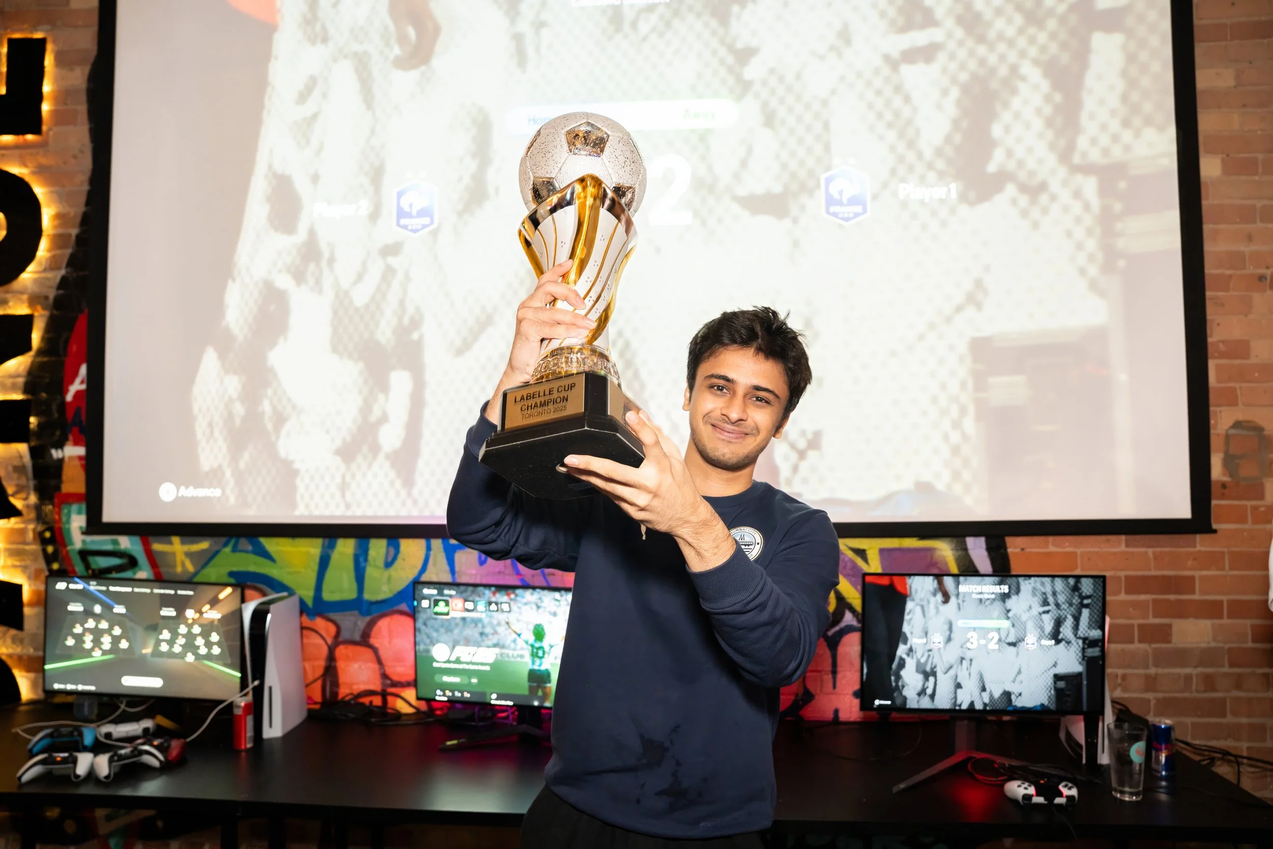 A young man holding a trophy that reads 'Labellé Cup Champion Toronto 2023,' smiling in front of a backdrop with a large screen displaying a sports video game, with gaming monitors and controllers on a desk underneath.