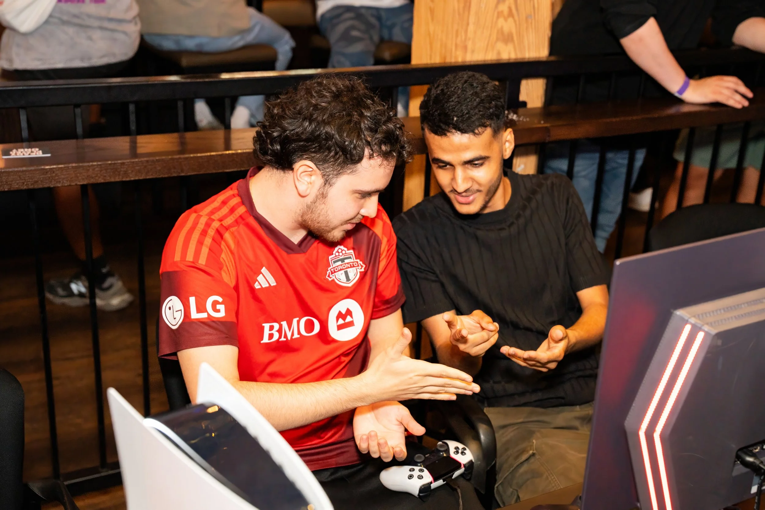 Two young men sitting at a gaming station, one wearing a red Toronto FC soccer jersey, both engaged in a conversation, with gaming controllers and a large monitor in front of them.