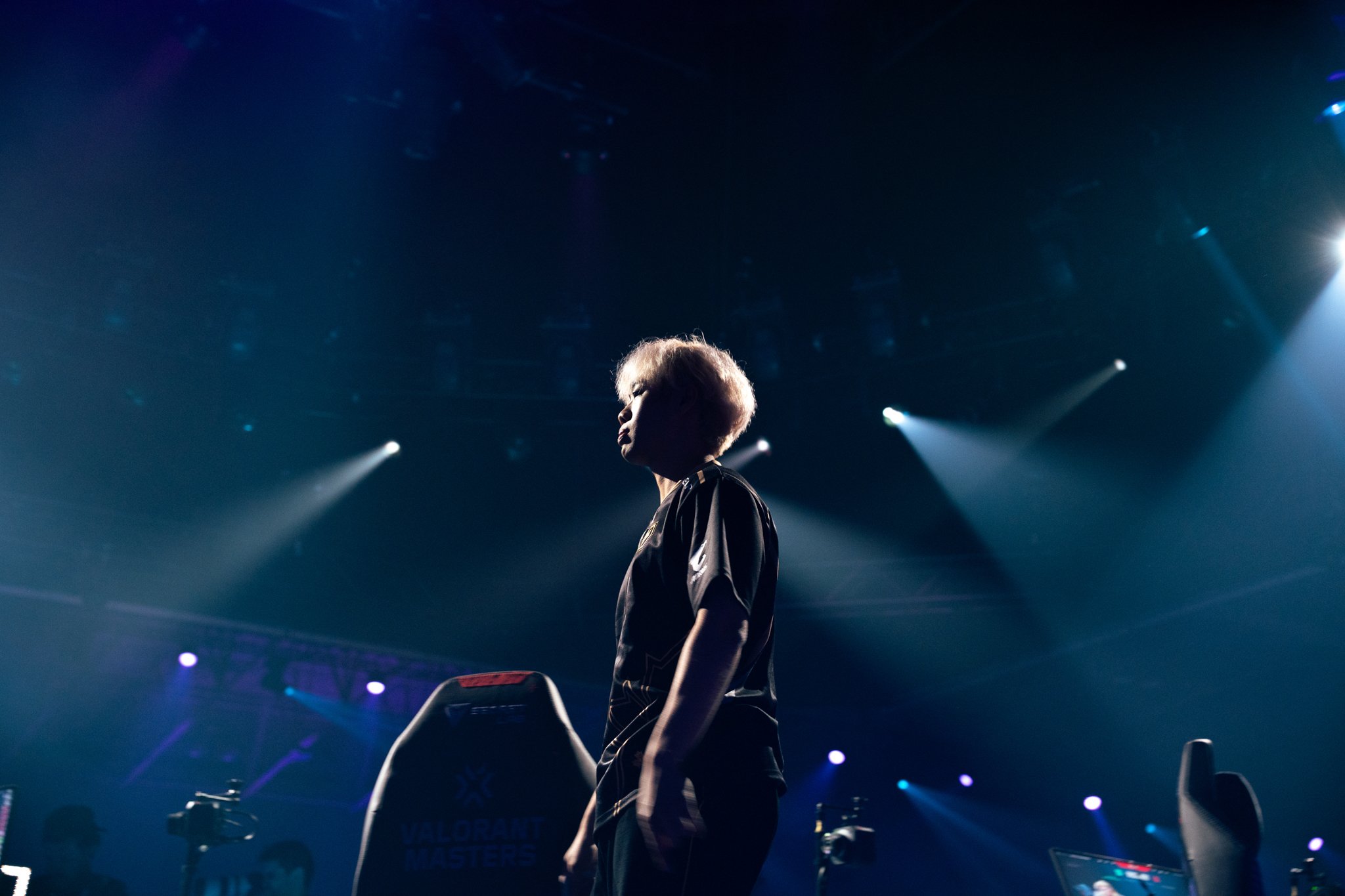 A person standing under bright stage lights at an esports event, with gaming chairs around them and a large screen displaying game graphics in the background.