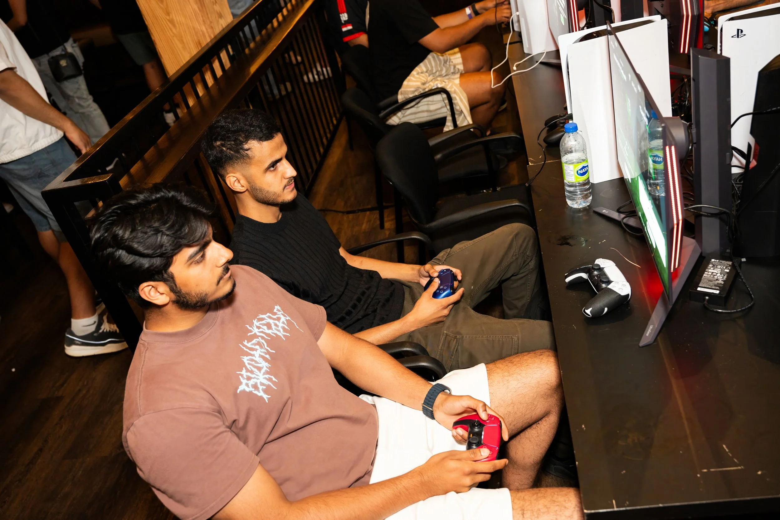 Two young men playing video games on a black desk in a gaming café, with monitors, controllers, and water bottles in front of them.