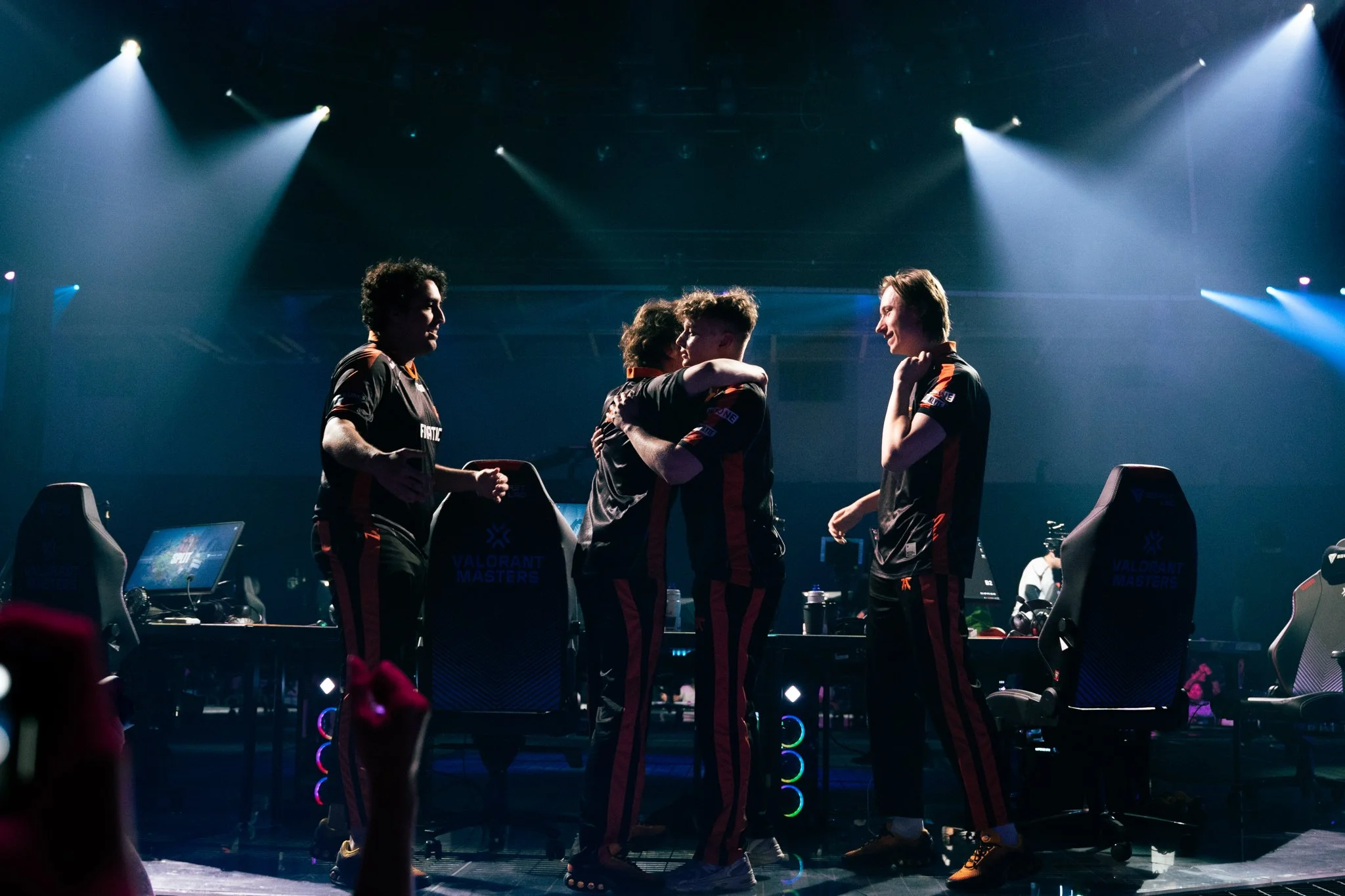 Group of esports players on stage, with two embracing and others smiling, surrounded by gaming chairs and monitors, under blue stage lighting.
