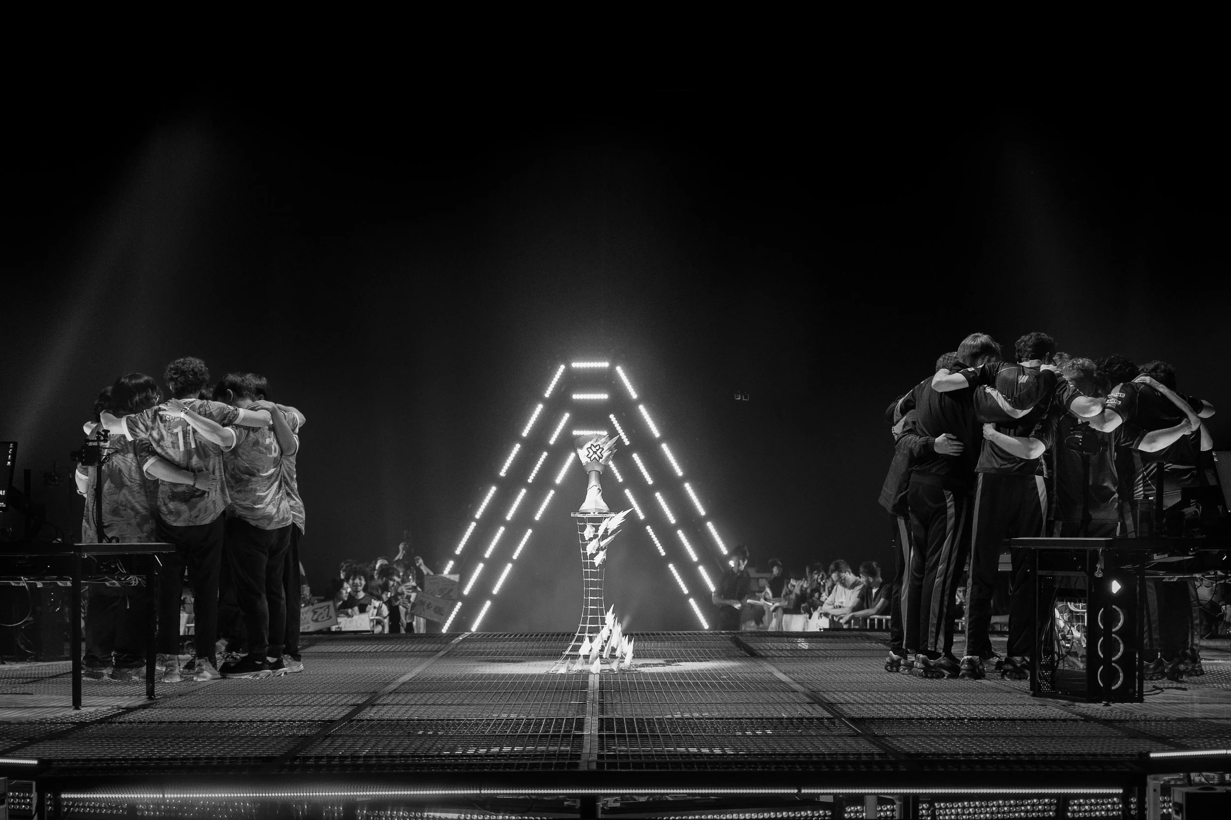 Black and white photo of two teams of esports players hugging on stage, with a trophy in the center and a crowd in the background.