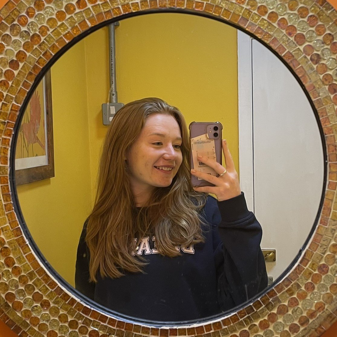 A young woman with long ginger hair taking a mirror selfie with her phone, smiling, in a room with yellow walls.