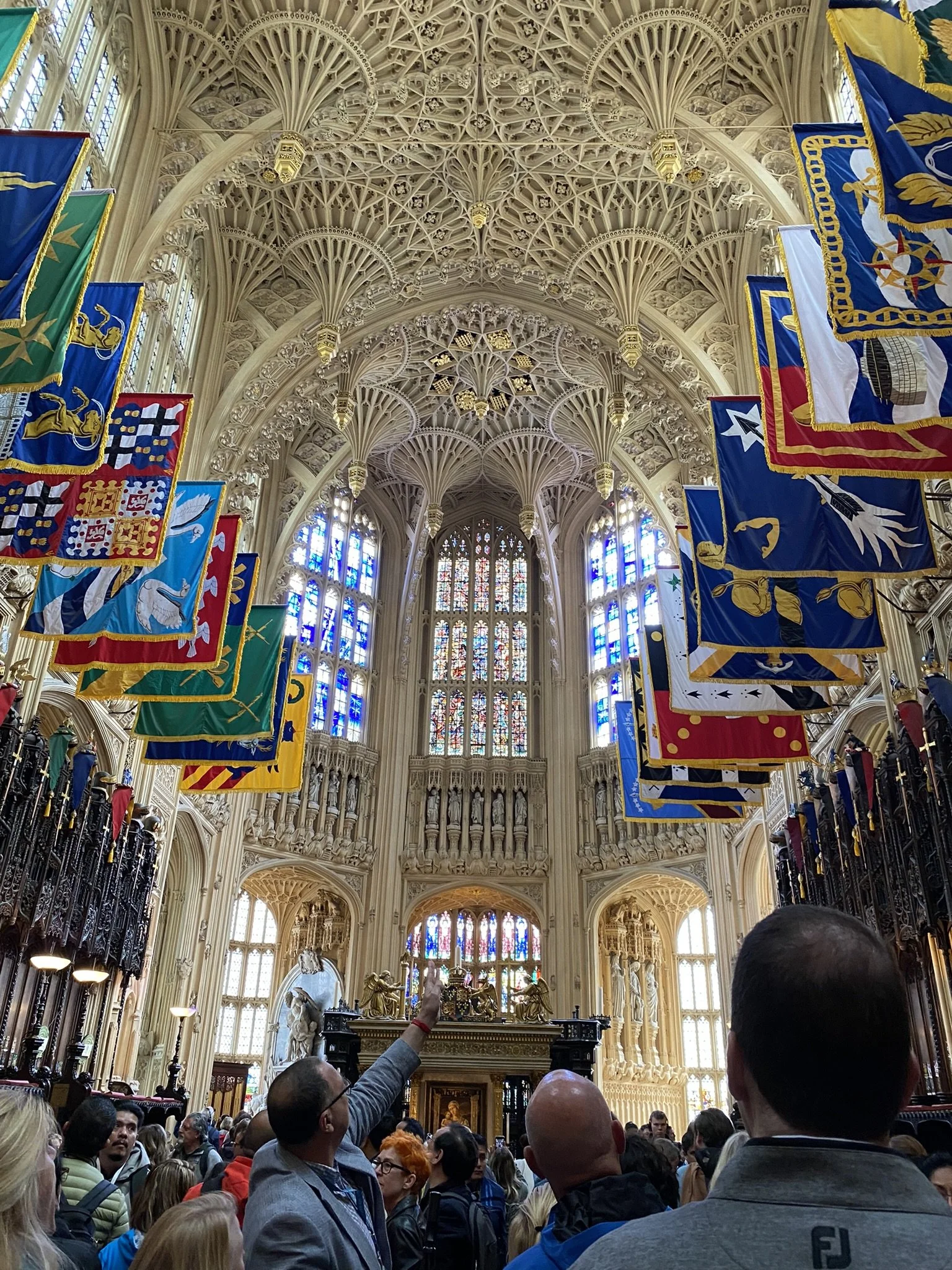Interior of a historic church or cathedral with intricate vaulted ceiling, stained glass windows, and banners hanging from the ceiling, with people gathered inside.