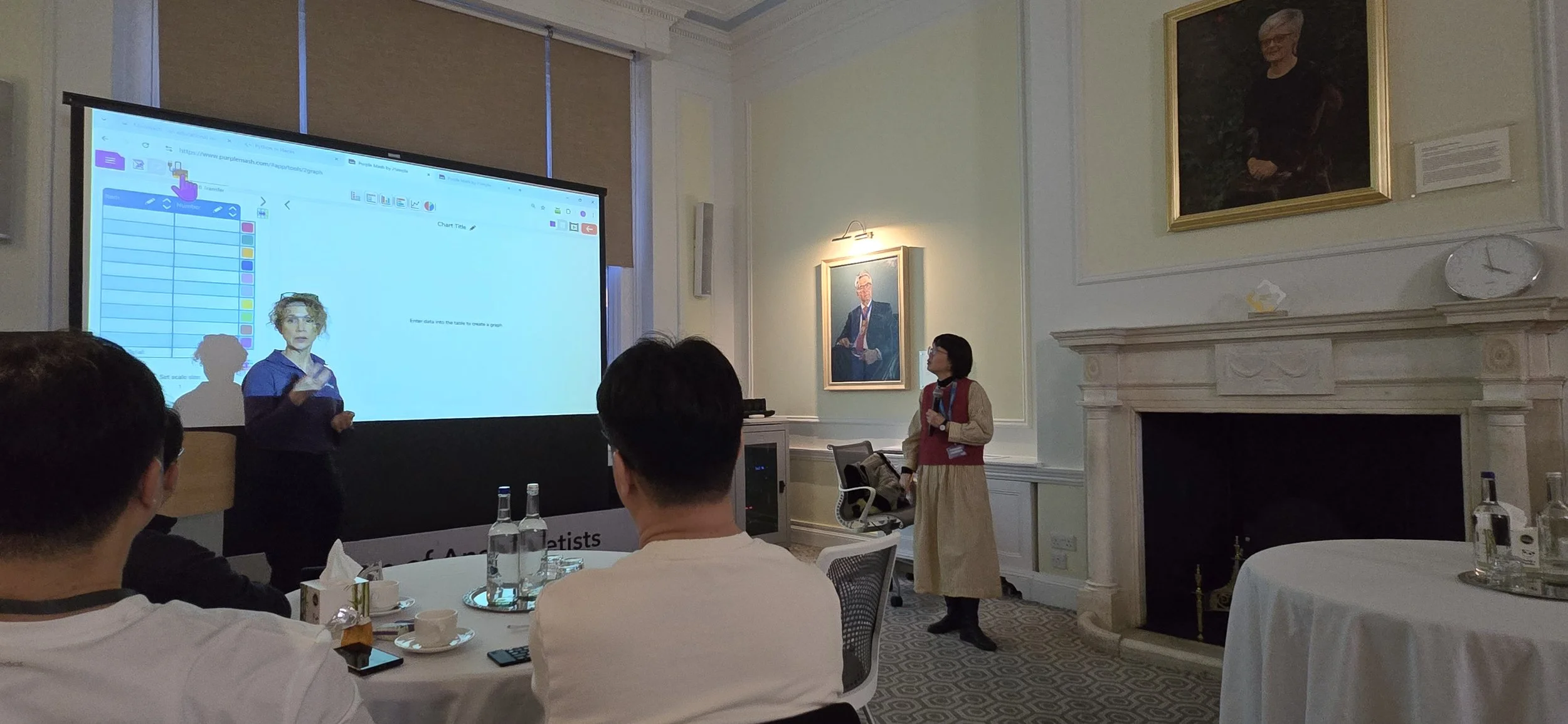 A woman presenting in a conference room with a large screen displaying a digital whiteboard, and an audience seated at tables with water bottles. Two framed portraits hang on the walls, and there is a fireplace in the room.