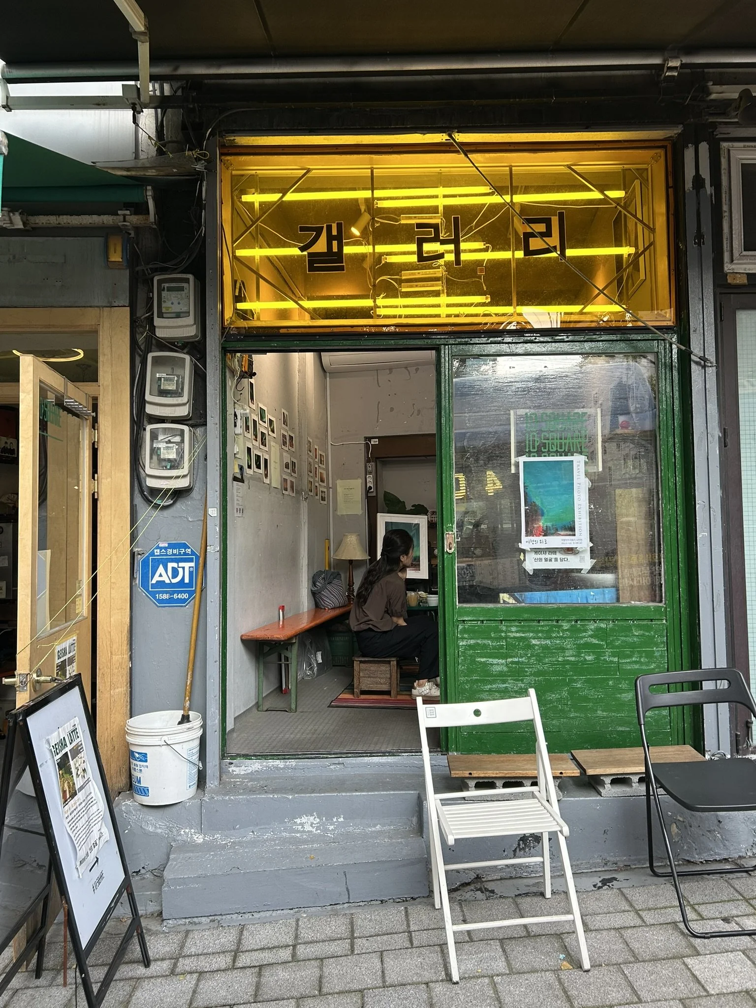A small Korean café with a neon sign in Hangul characters, a woman sitting inside, and outdoor chairs in front.