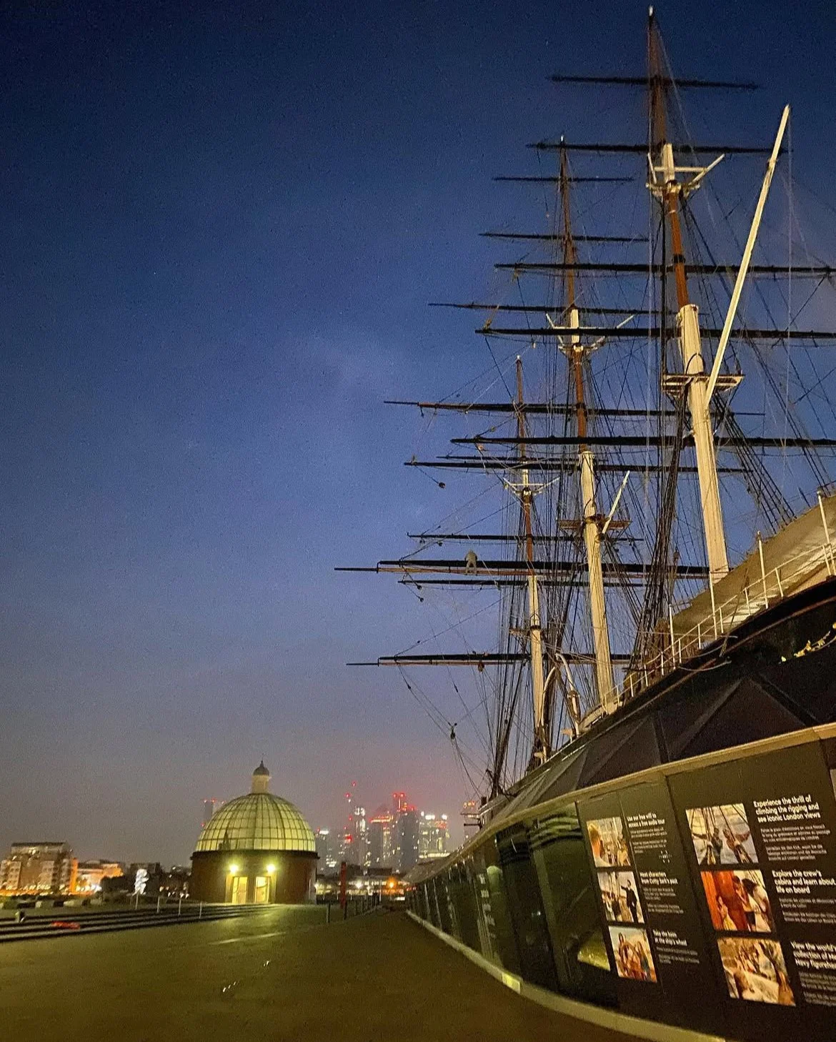 Night view of a historic sailing ship with tall masts, docked on a city waterfront, with a domed building and city skyline illuminated in the background.