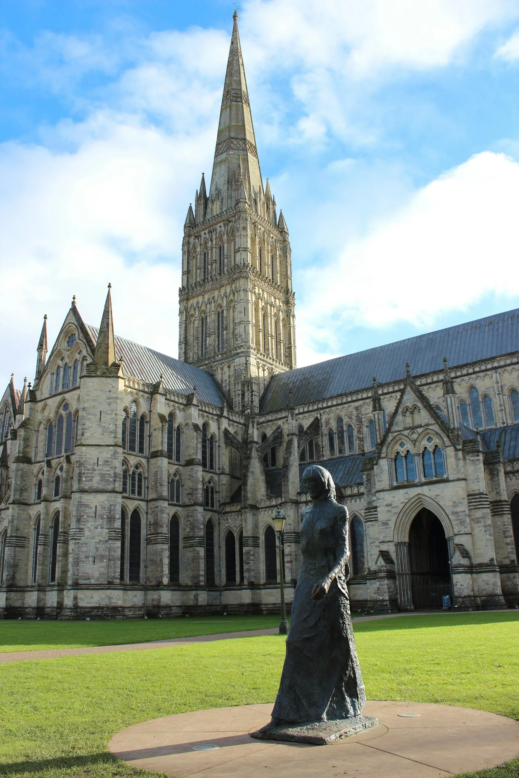 A historic cathedral with a tall steeple, stone walls, and Gothic architecture, with a bronze sculpture of a woman in a dress in the foreground on a grassy lawn under a blue sky.
