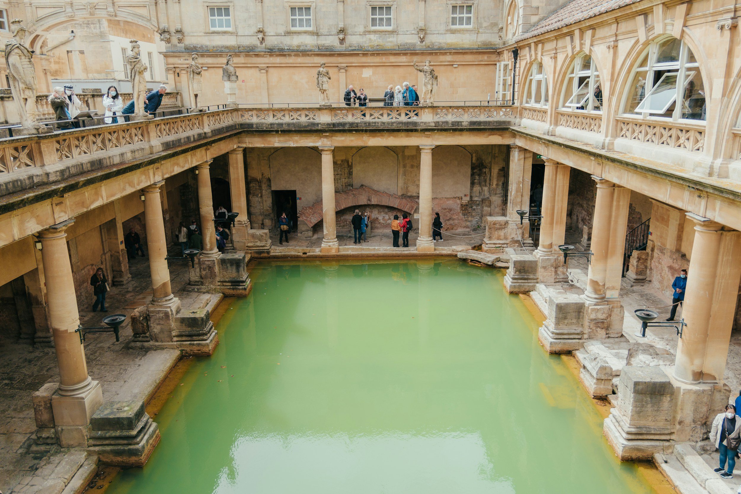 Ancient Roman cistern with a rectangular pool of green water, surrounded by stone columns, arches, and statues, with visitors exploring the site.