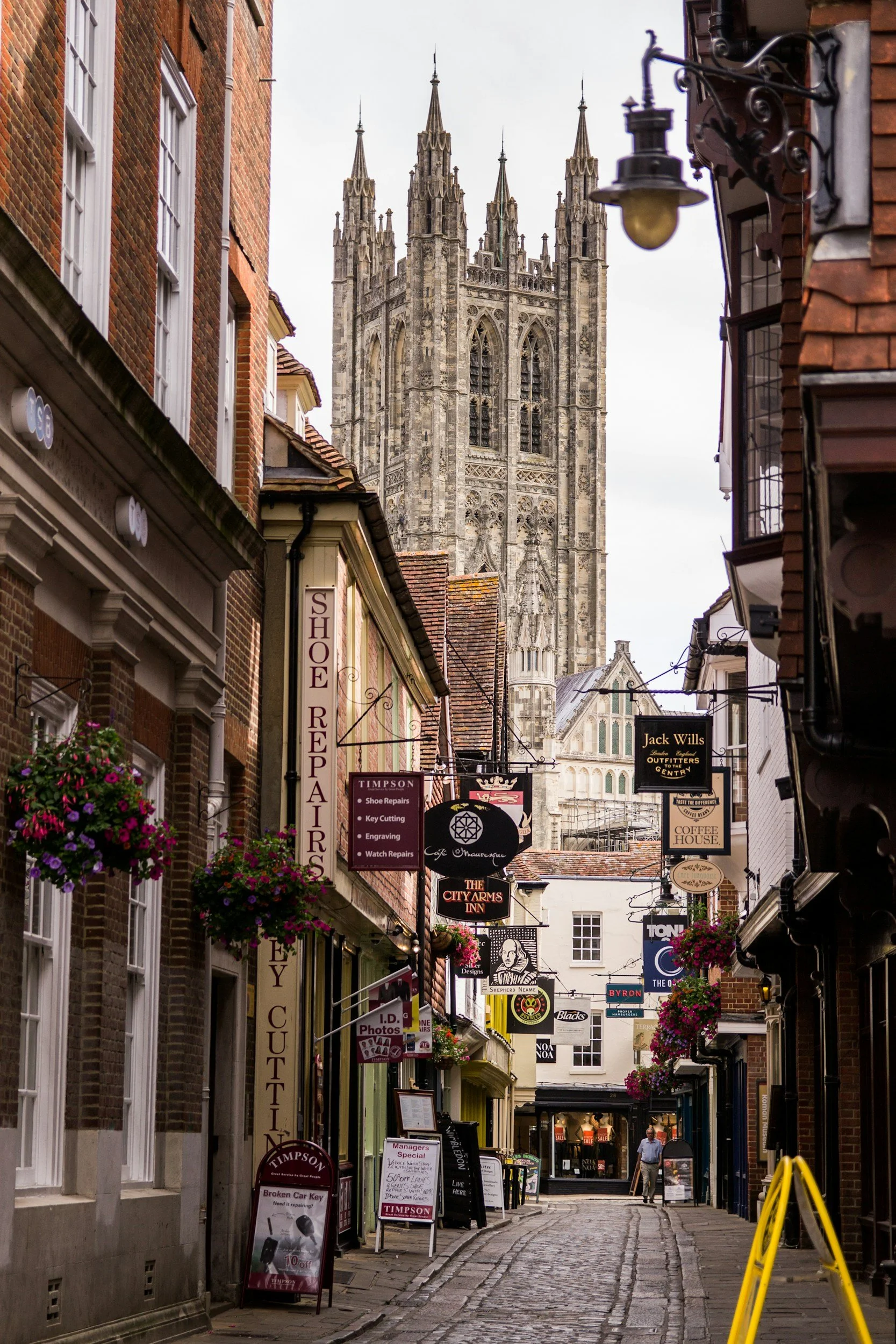 A narrow cobblestone street lined with shops and hanging flower baskets, with a large Gothic-style cathedral tower in the background.