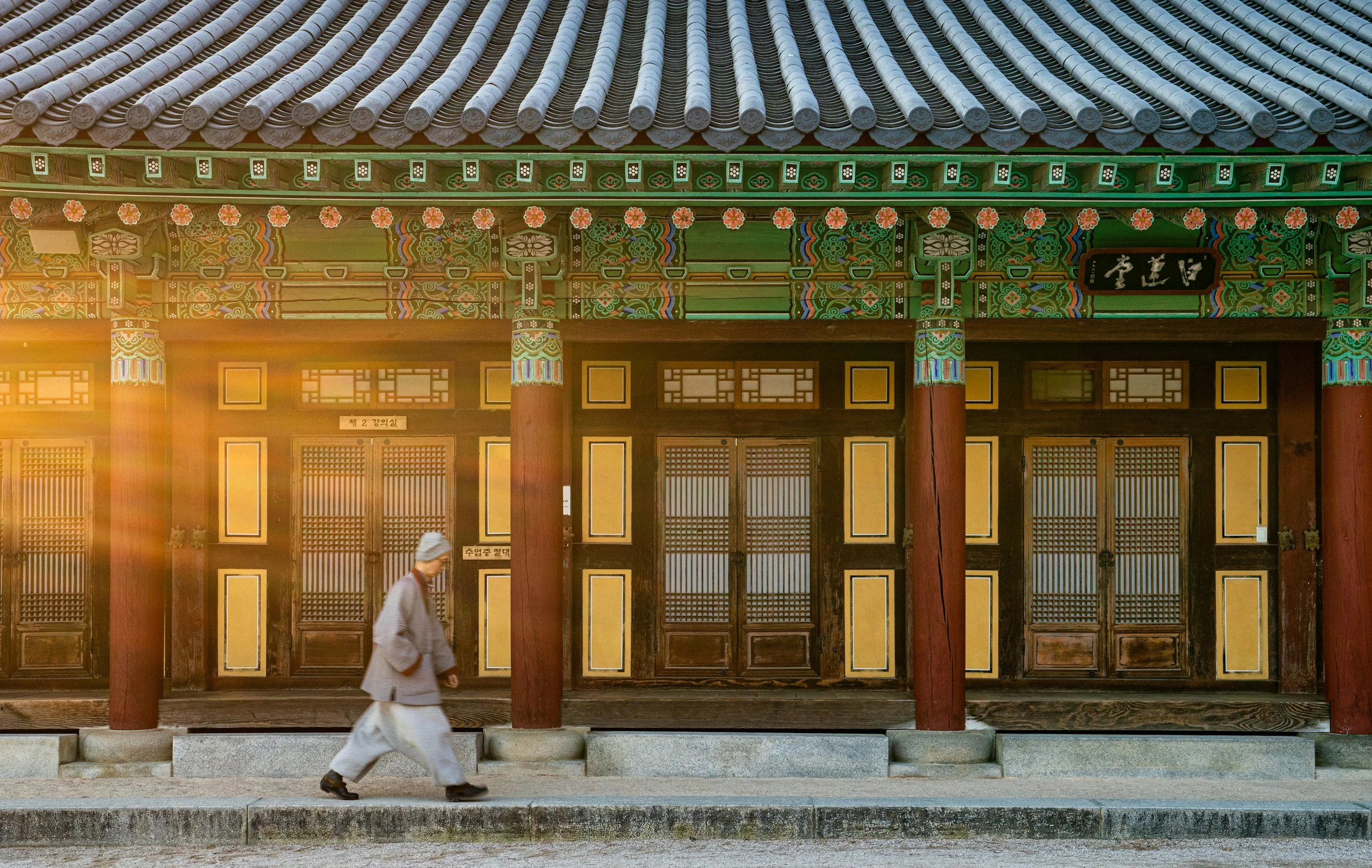 A man in traditional clothing walking past a colorful historic Korean temple with ornate roof and wooden doors.