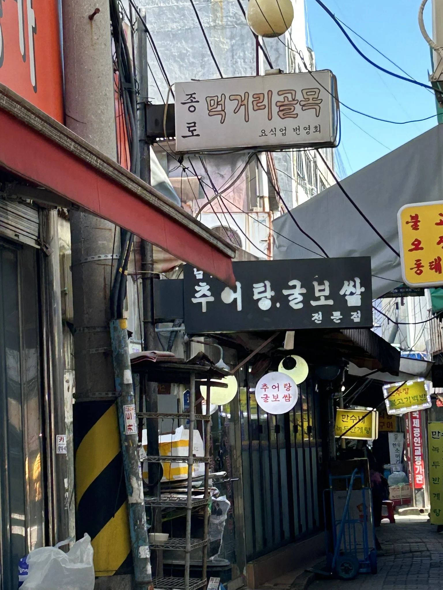 A busy street in Korea with various signs in Korean, including a sign for a restaurant and other small businesses, along with utility poles, wires, and street vendors.