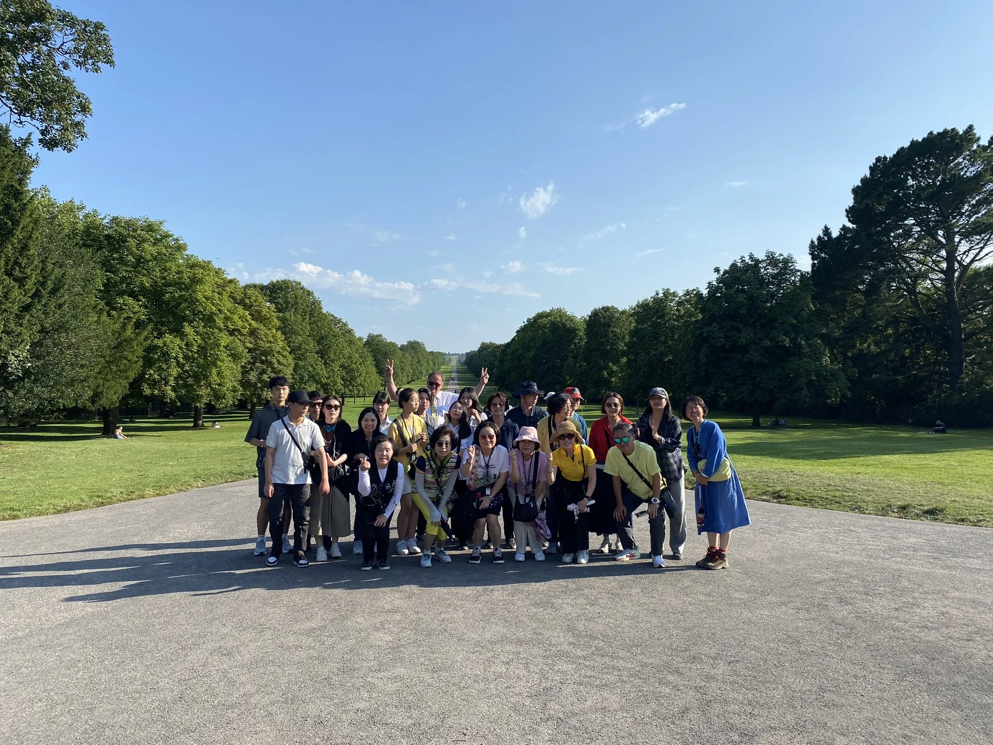 A large group of people posing for a photo outdoors on a sunny day, with a park and trees in the background.