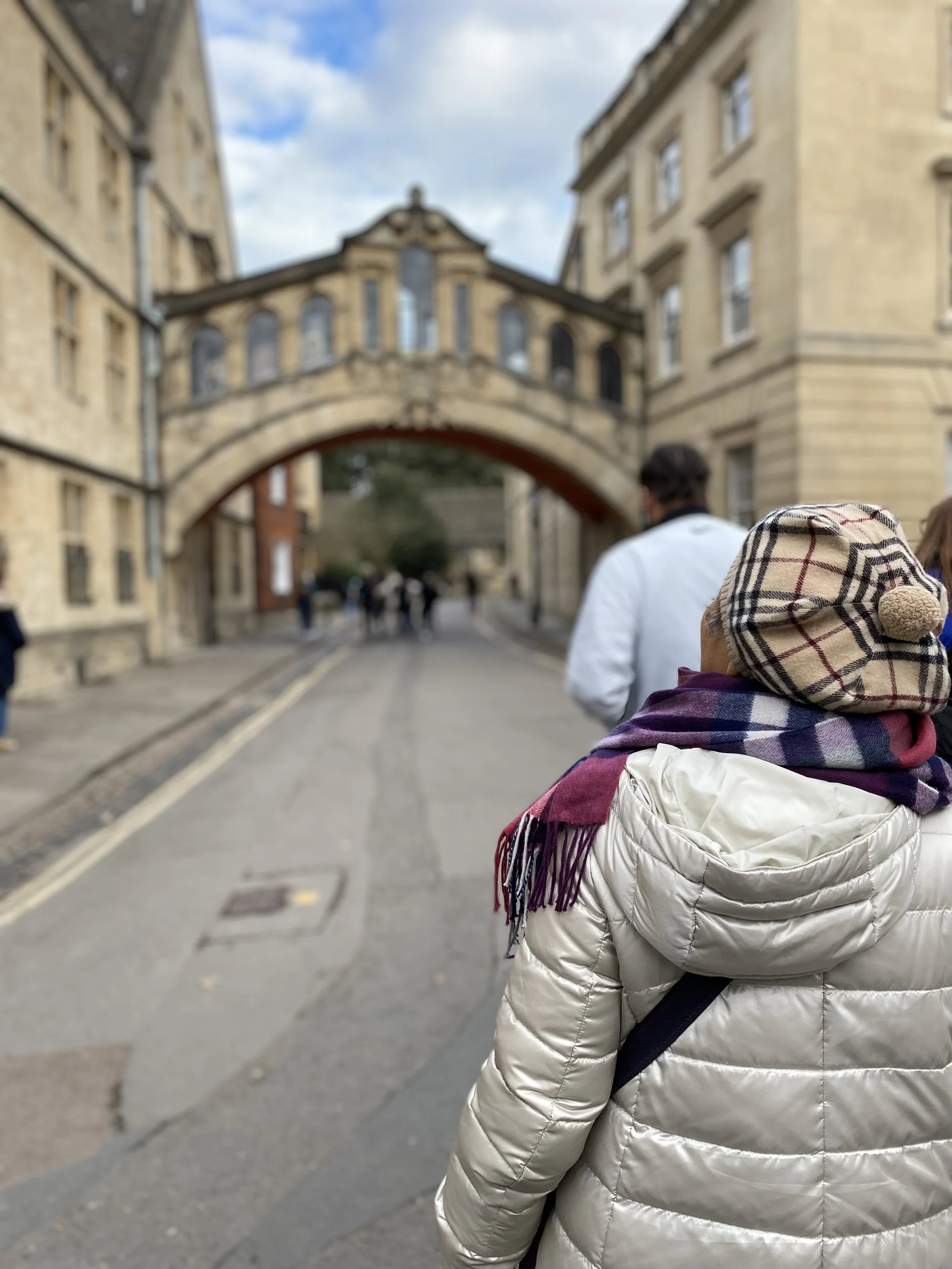 People walking on a street with historic buildings, a bridge connecting the buildings, and a cloudy sky.