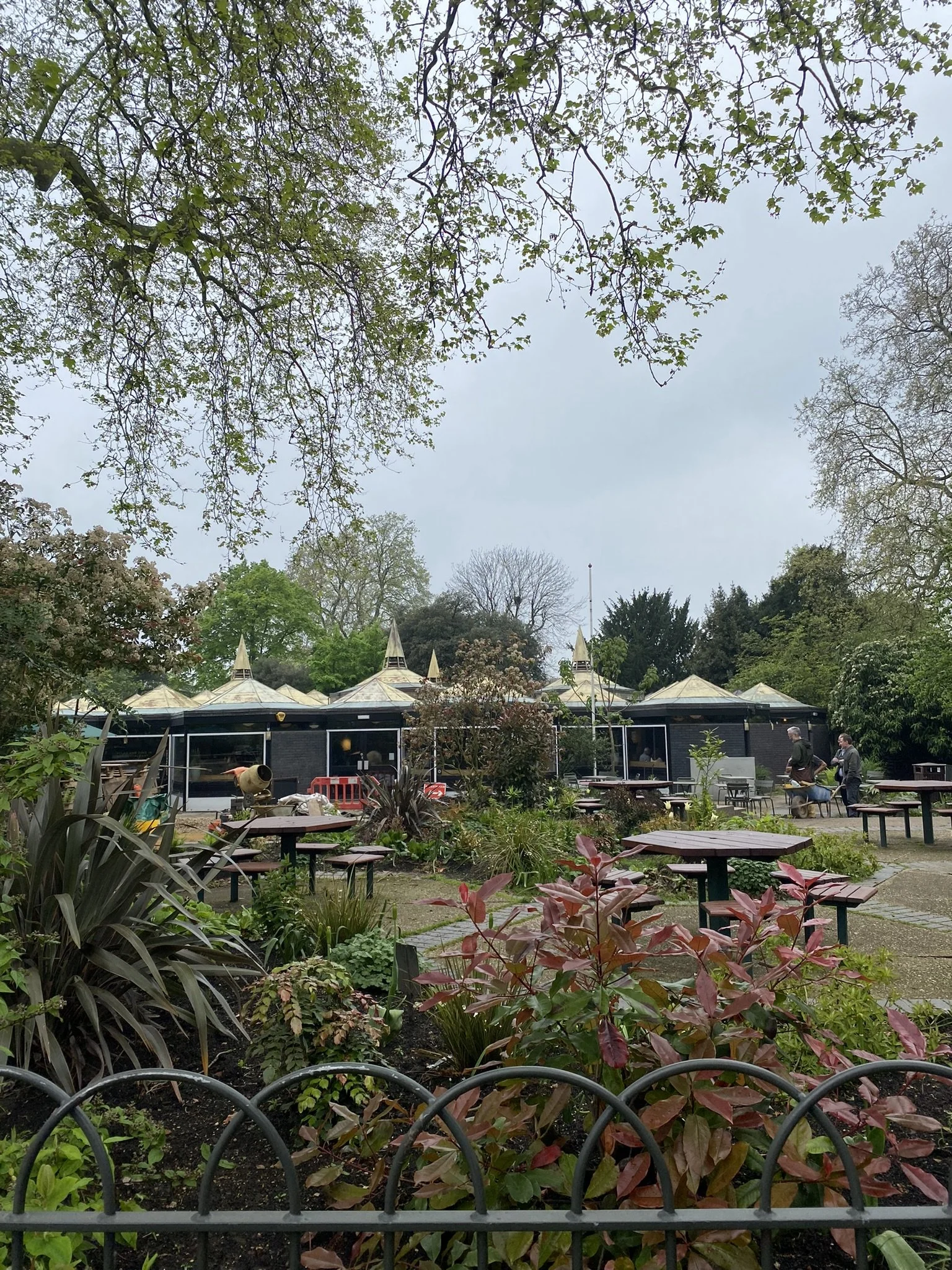 Image of an outdoor garden with green plants, patio tables, and people walking in the background on a cloudy day.