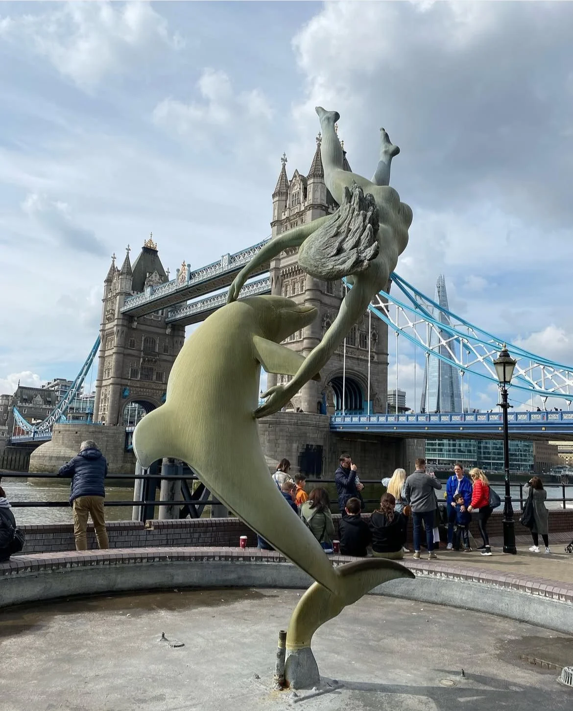 Sculpture of a mermaid holding a trident with City Hall and Tower Bridge in the background, near the River Thames in London.