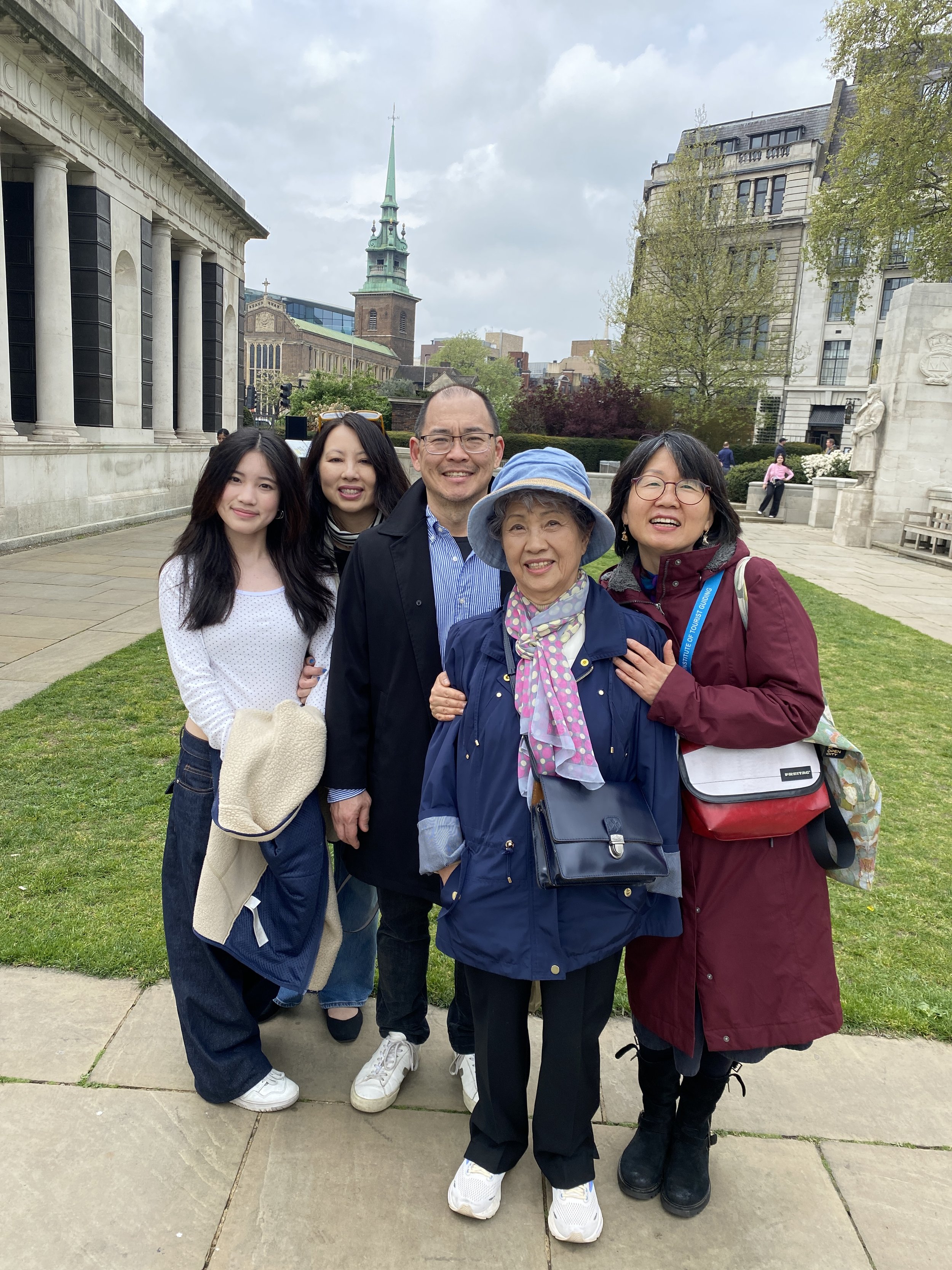 Group of five people standing on a sidewalk in front of historic buildings and a church in an urban park setting, with cloudy skies overhead.