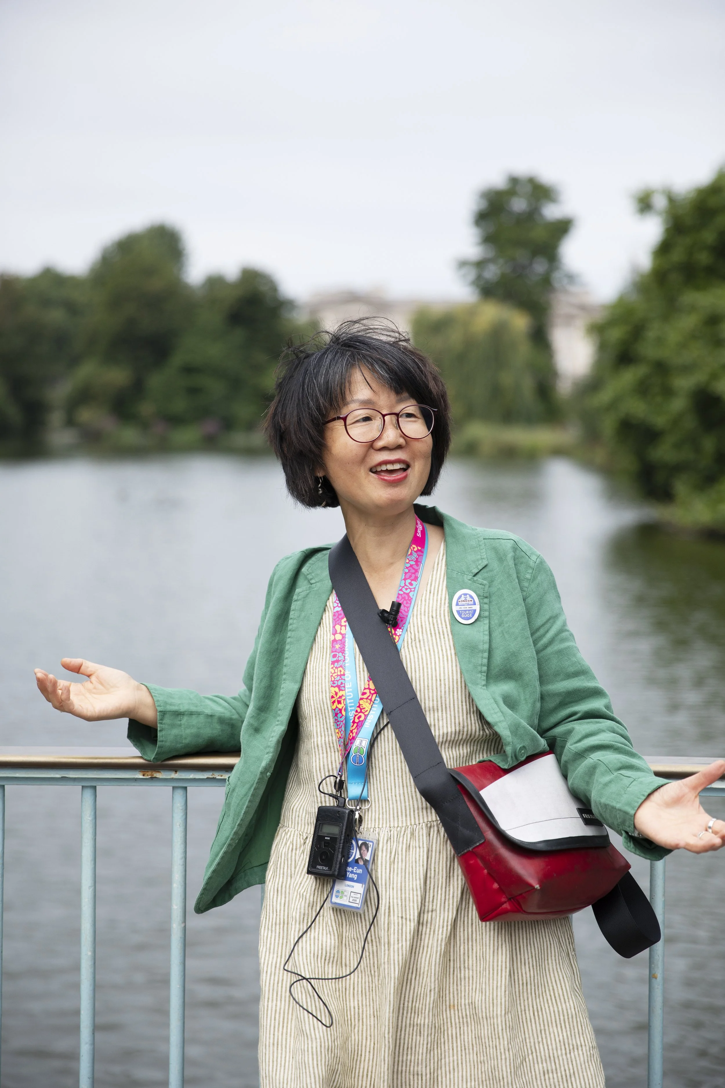 Woman with glasses, short dark hair, green blazer, and beige dress standing by a river, speaking or explaining with arms open outdoors.