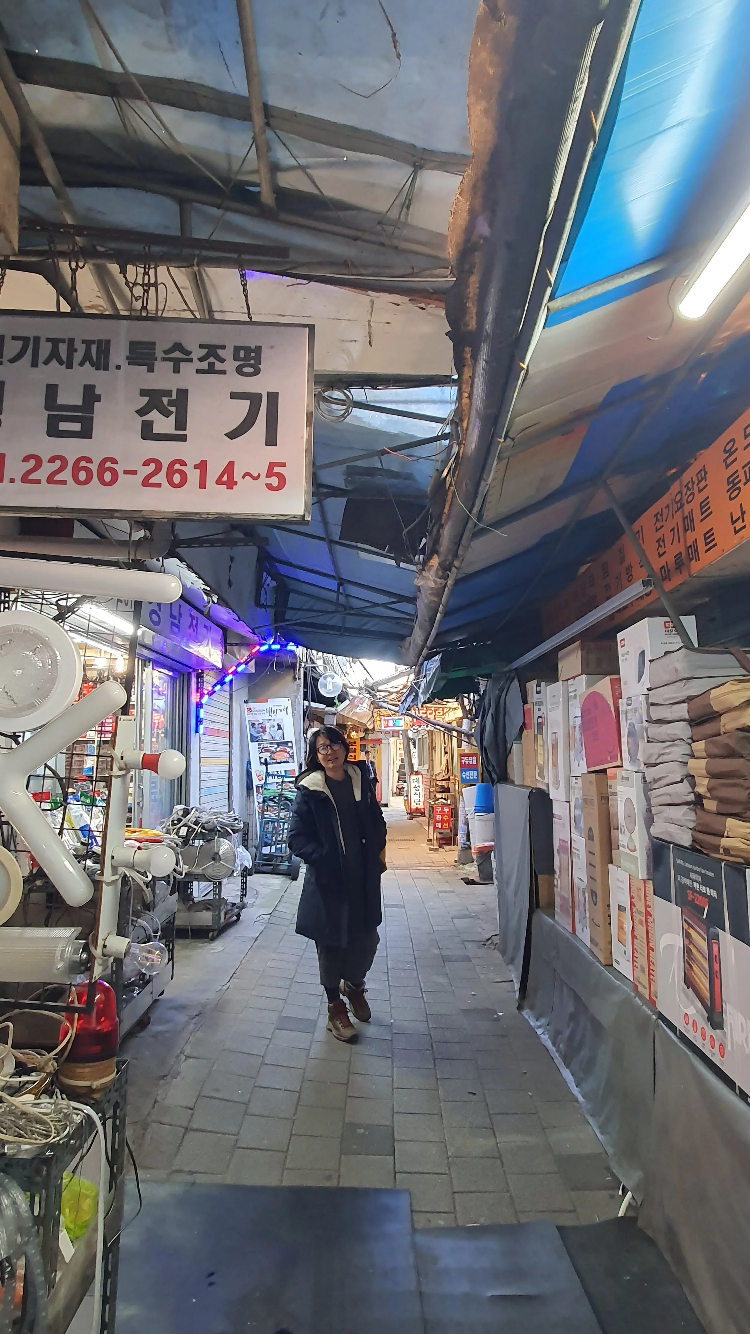 A woman walking through a narrow outdoor market alley lined with various shops and electronic items, with overhead signs and colorful lights, possibly in Korea.
