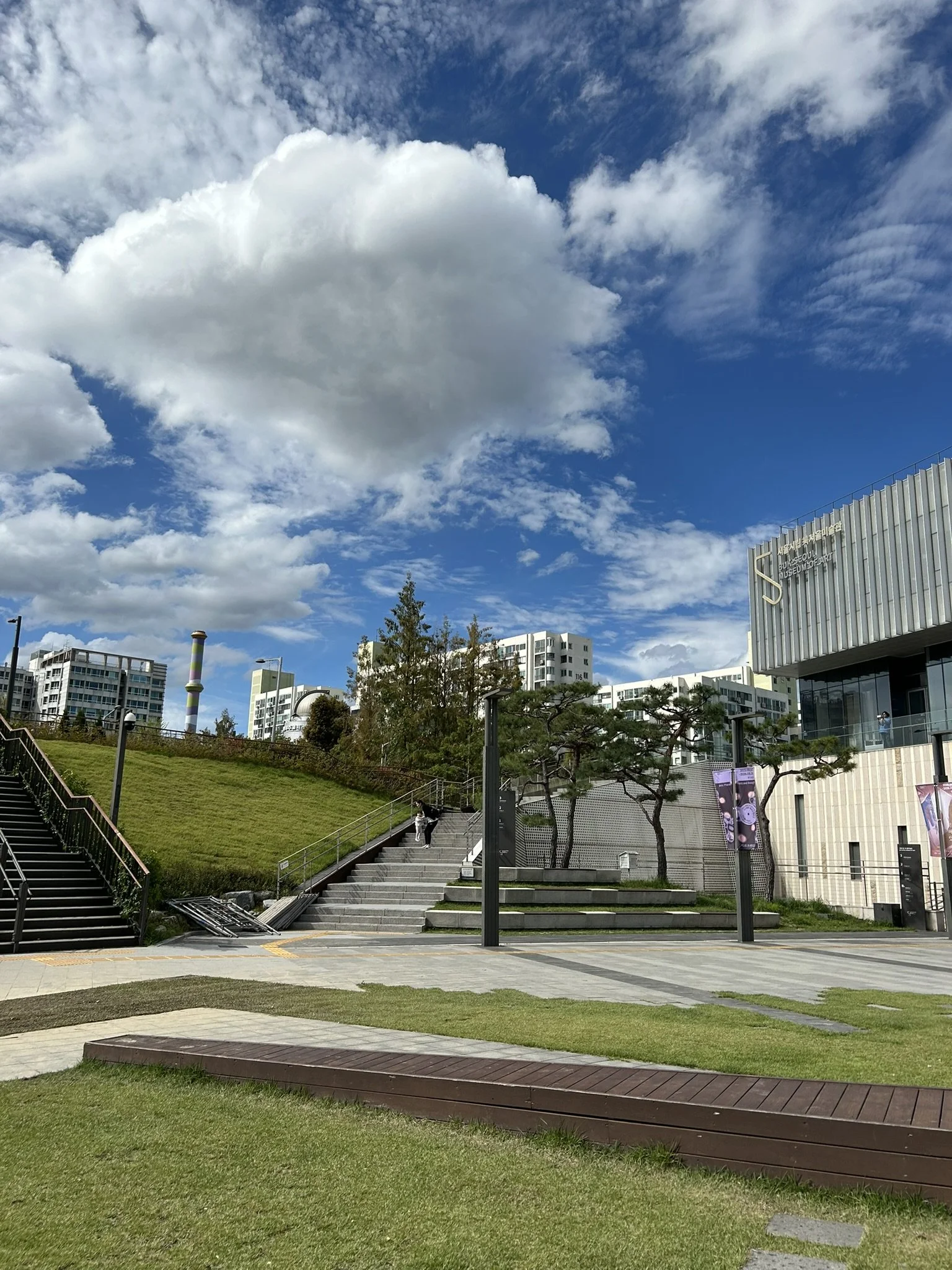 Urban park with stairs, grass, trees, modern buildings, and a partly cloudy sky.