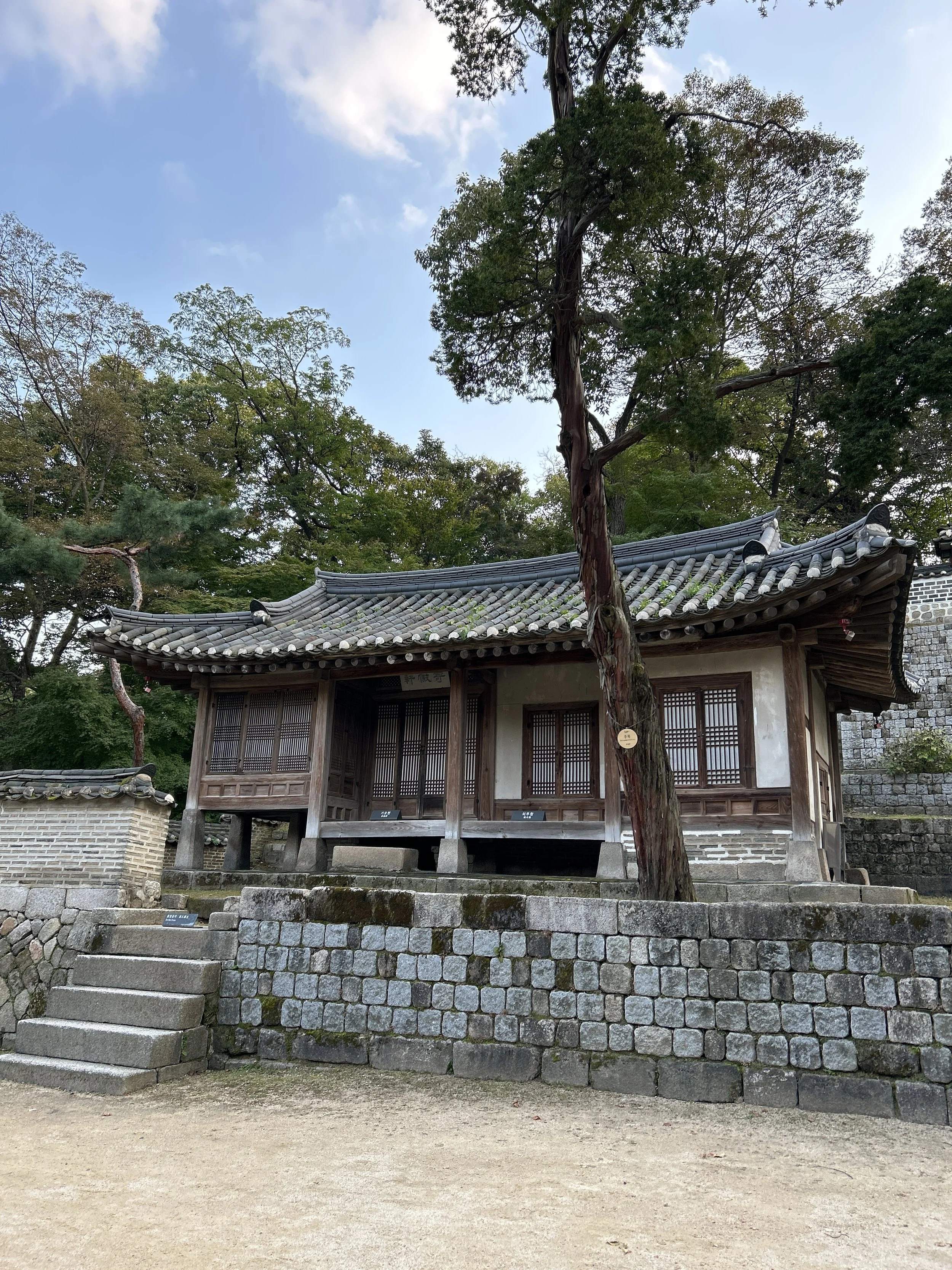 A traditional Korean wooden building with a tiled roof, elevated on stone platforms, surrounded by trees and greenery, under a partly cloudy sky.