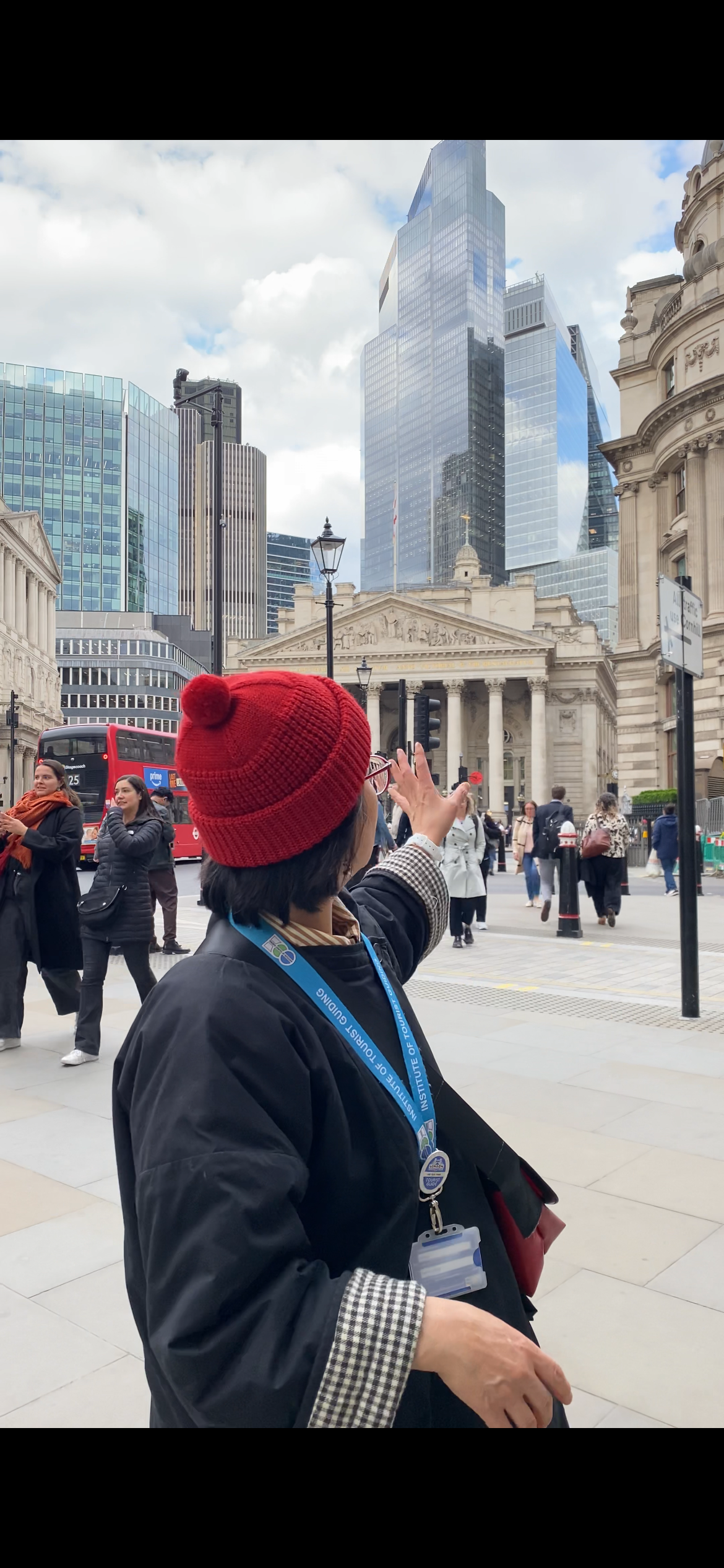 A woman with a red beanie and glasses pointing towards the cityscape, with tall skyscrapers and historic buildings in the background, in a busy urban area.