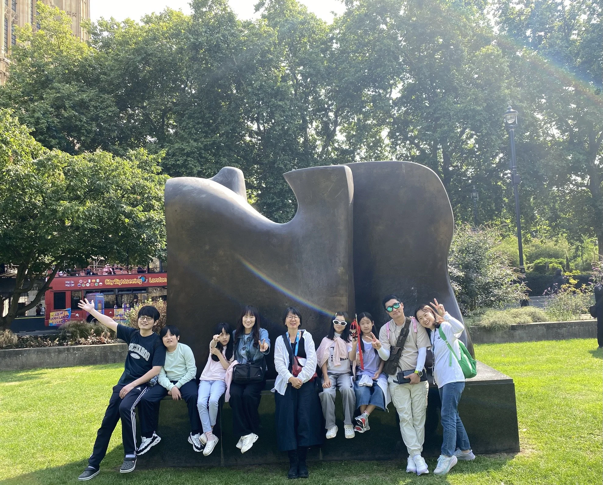 Group of people sitting and standing in a park in front of a large abstract black sculpture, with trees and a red double-decker bus in the background.
