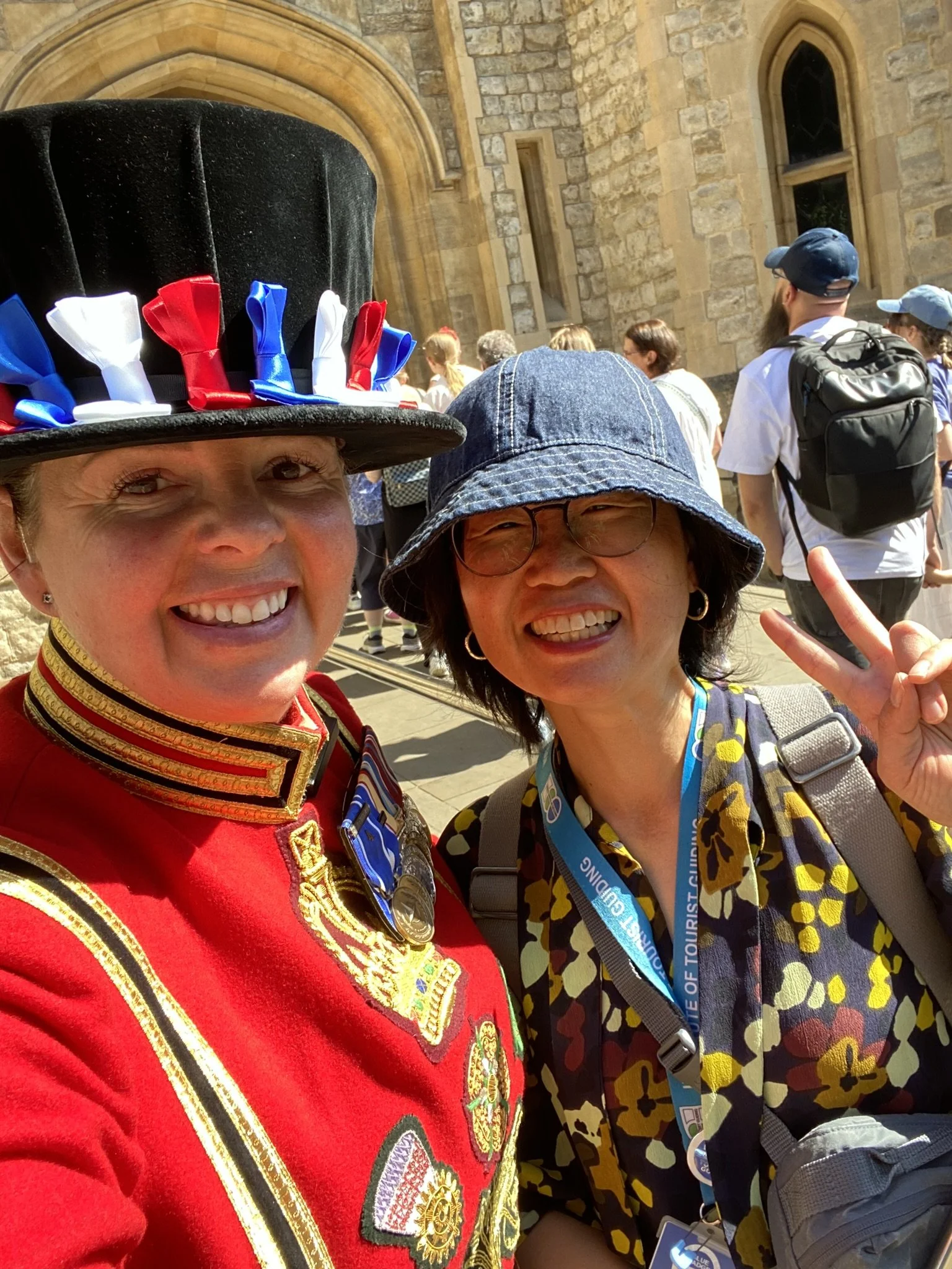 Two women smiling for a selfie outdoors. The woman on the left is dressed as a marching band member with a tall black hat decorated with red, white, and blue ribbons, and a red uniform with gold details. The woman on the right is wearing a denim bucket hat, glasses, a floral shirt, and a lanyard, and is making a peace sign with her hand.