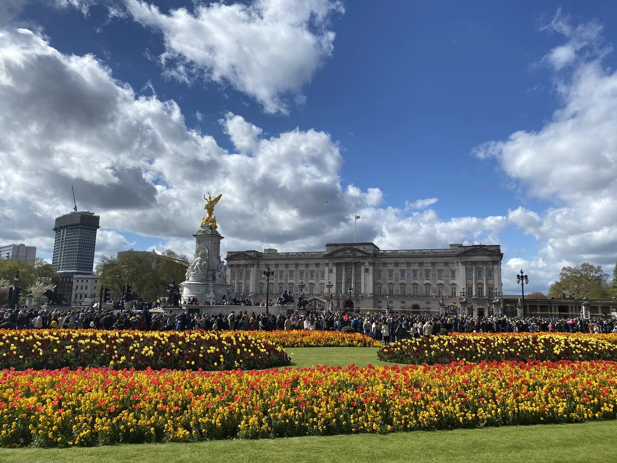 Buckingham Palace with a large crowd of visitors, a golden statue, colorful flowerbeds, and a partly cloudy sky.