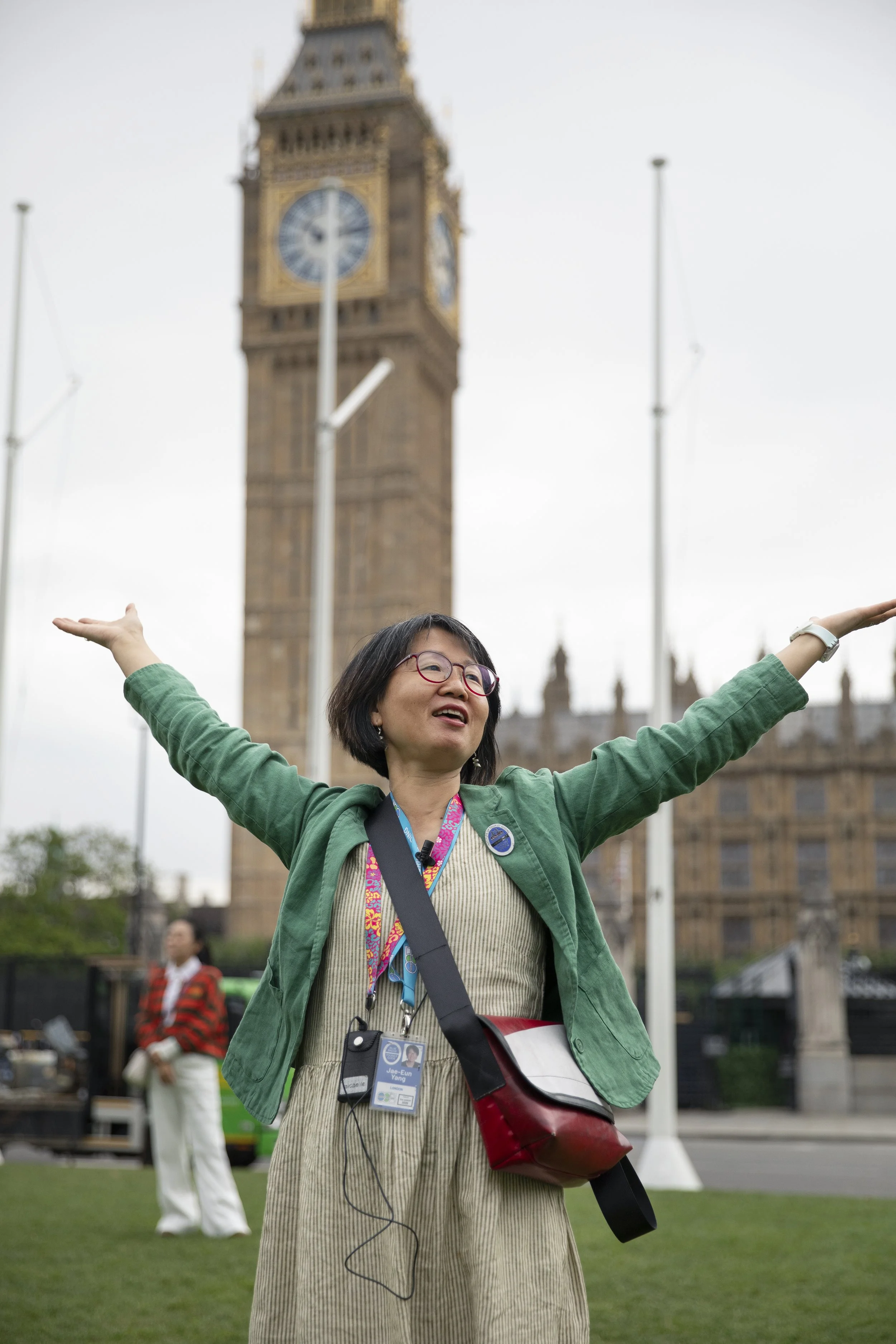 A woman standing outdoors with arms outstretched in front of Big Ben, London. She is smiling, wearing glasses, a green jacket, a beige dress, and a badge around her neck.
In the background, another person dressed in red and white is visible, along with a green vehicle and some light poles.