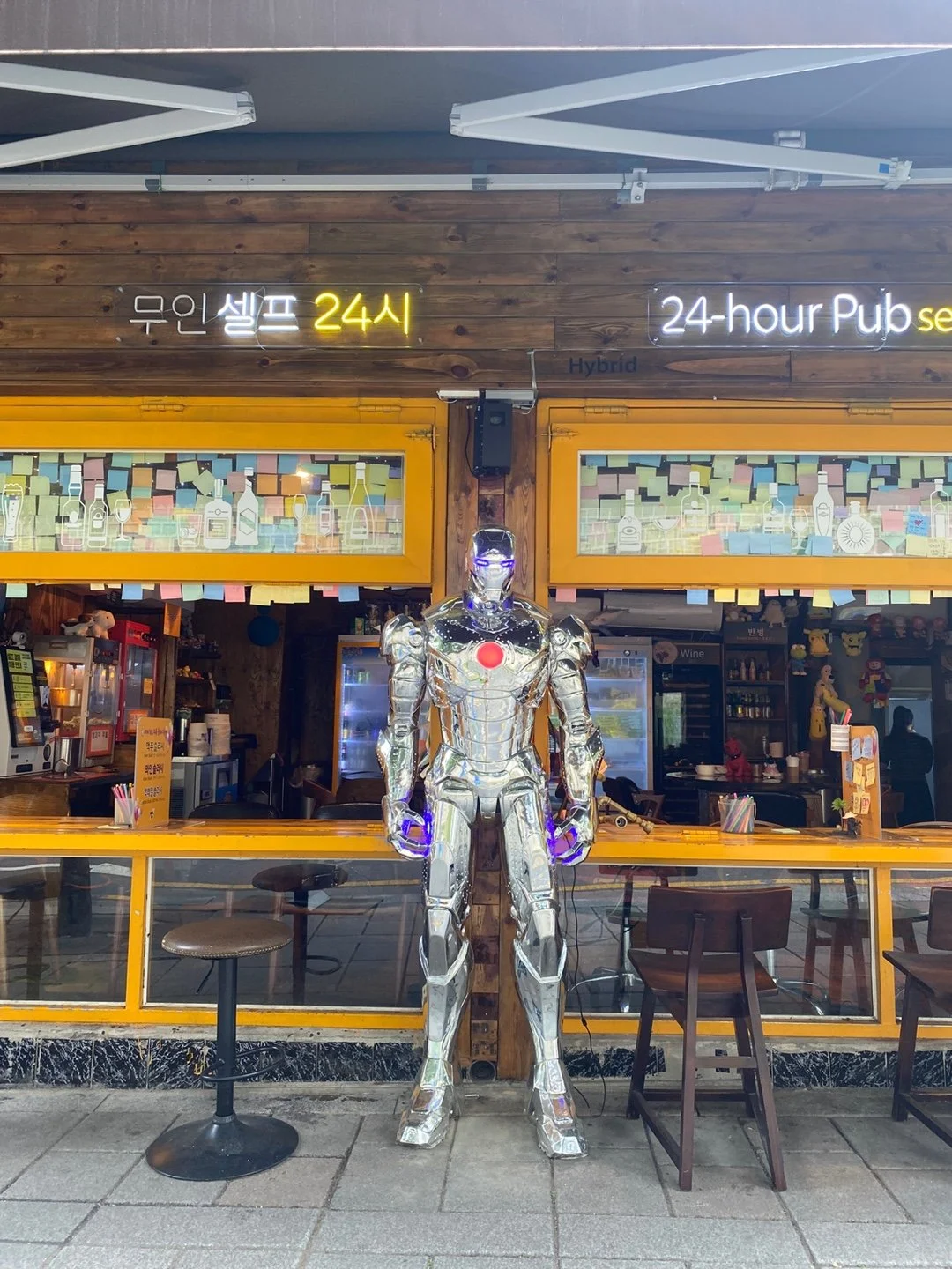 A shiny, robotic Iron Man figure standing outside a 24-hour pub, with a wooden and yellow exterior, neon signage, and colorful storefront decorations.