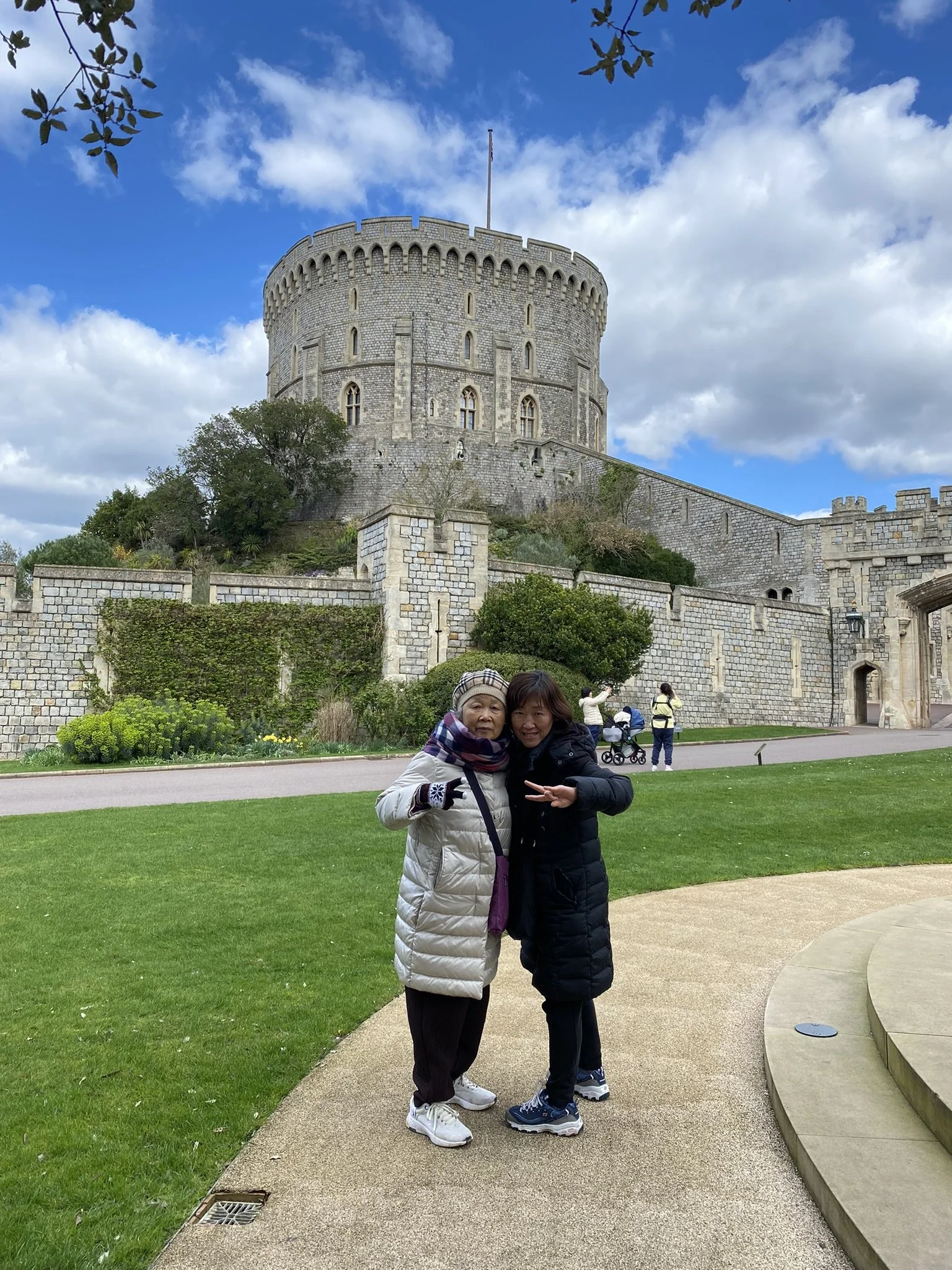 Two women standing together and smiling in front of a historic stone castle with a round tower, under a partly cloudy sky.