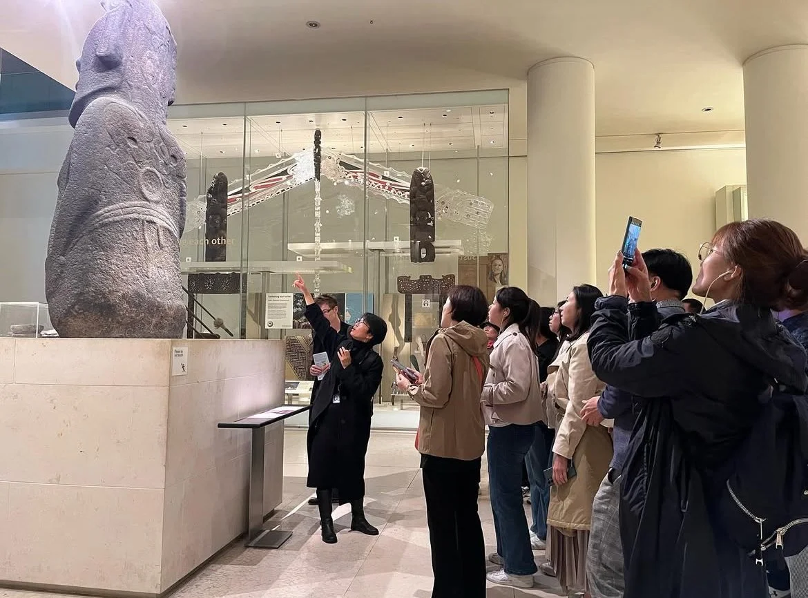 A group of people in a museum observing and taking pictures of ancient stone statues and artifacts displayed behind glass.