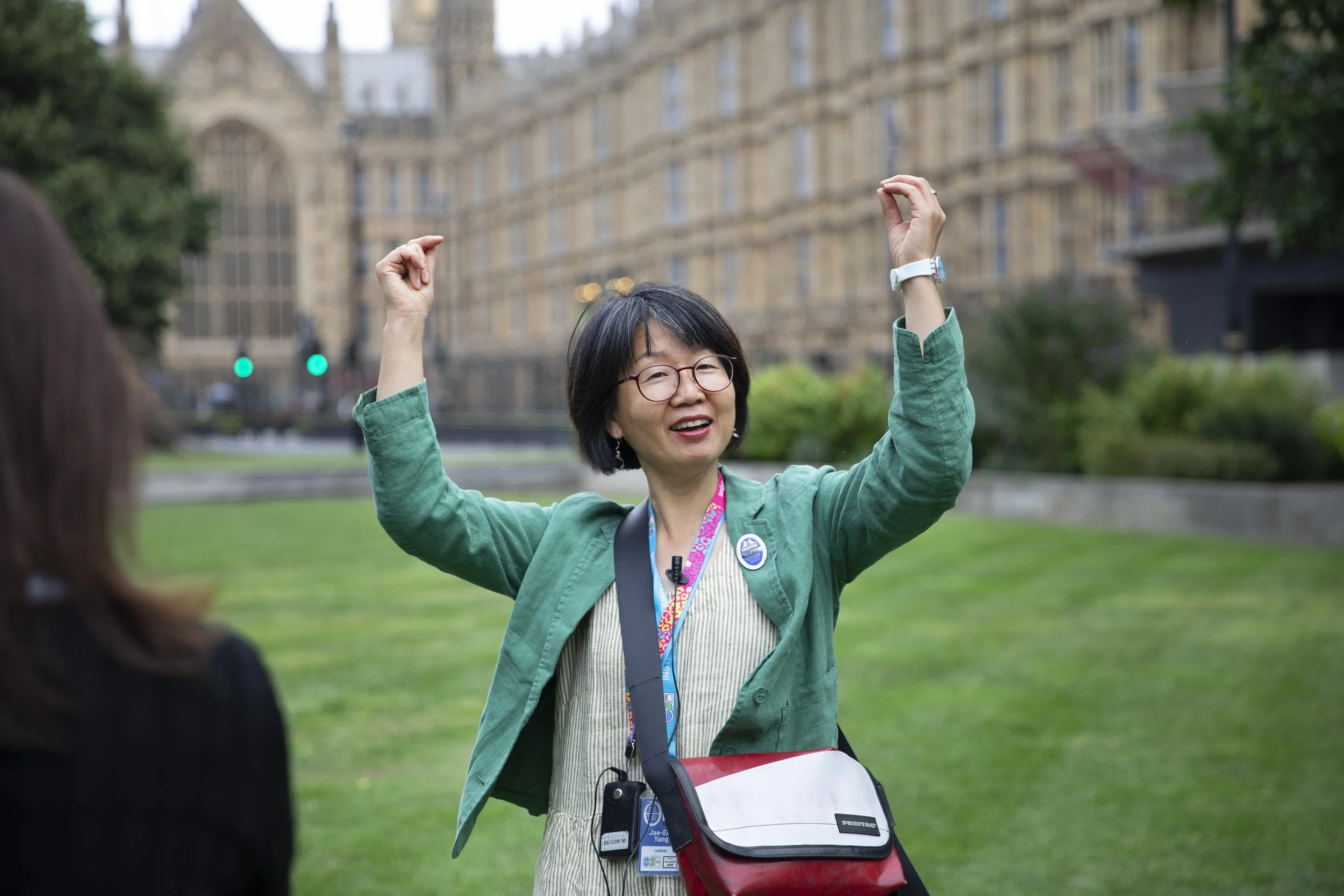 A woman with short black hair and glasses, smiling and raising her arms, standing outdoors in front of a historic building, wearing a green blazer, a lanyard, and a red bag.