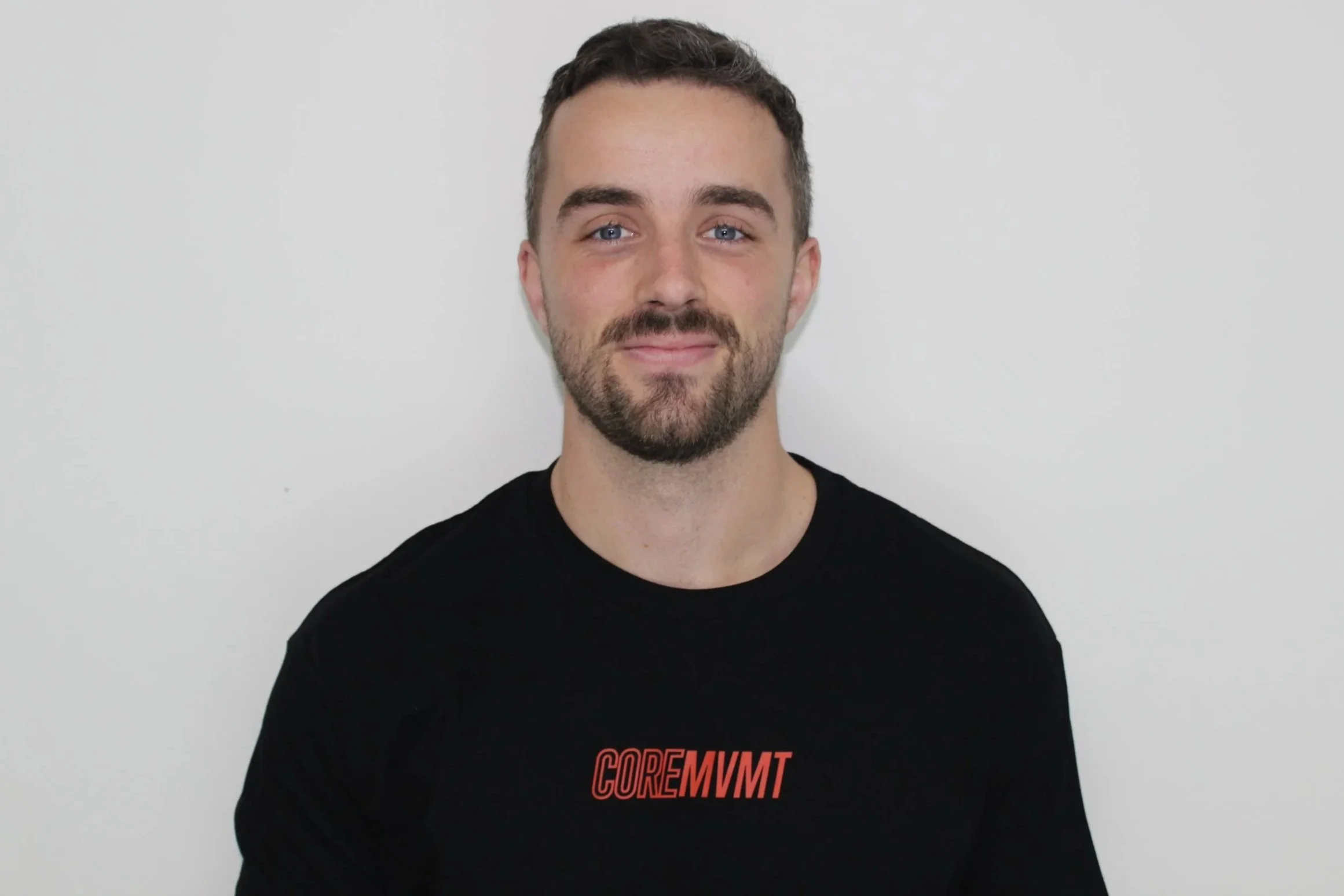 A young man with short dark hair, a beard, and blue eyes is smiling in front of a plain white background. He is wearing a black sweatshirt with the red text 'COREMVMT' on it.