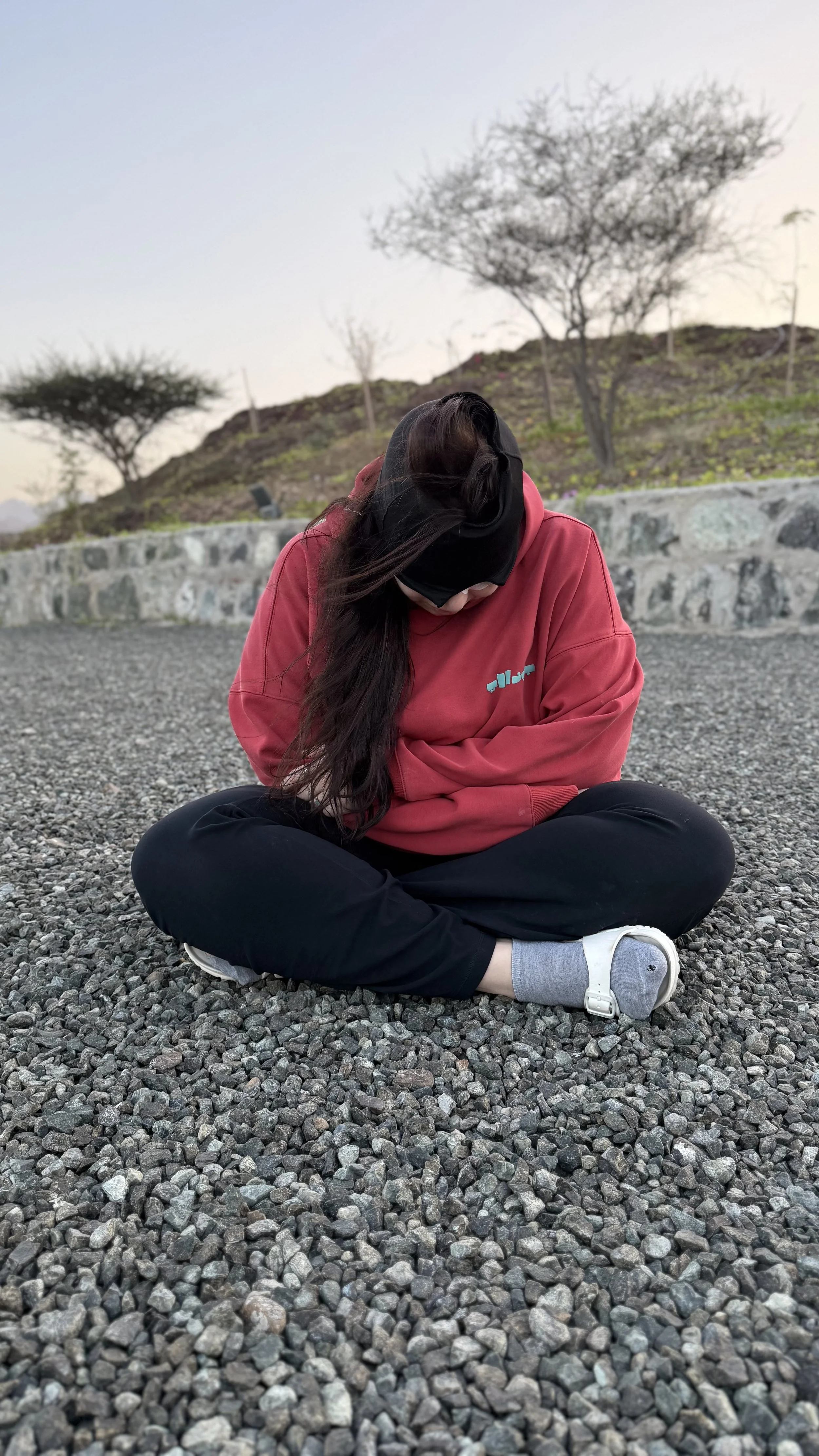 A person sitting cross-legged on a gravel surface, wearing a red hoodie, black pants, white shoes, and a black headband, looking down with long dark hair covering part of their face. In the background, there are some trees and a stone wall.