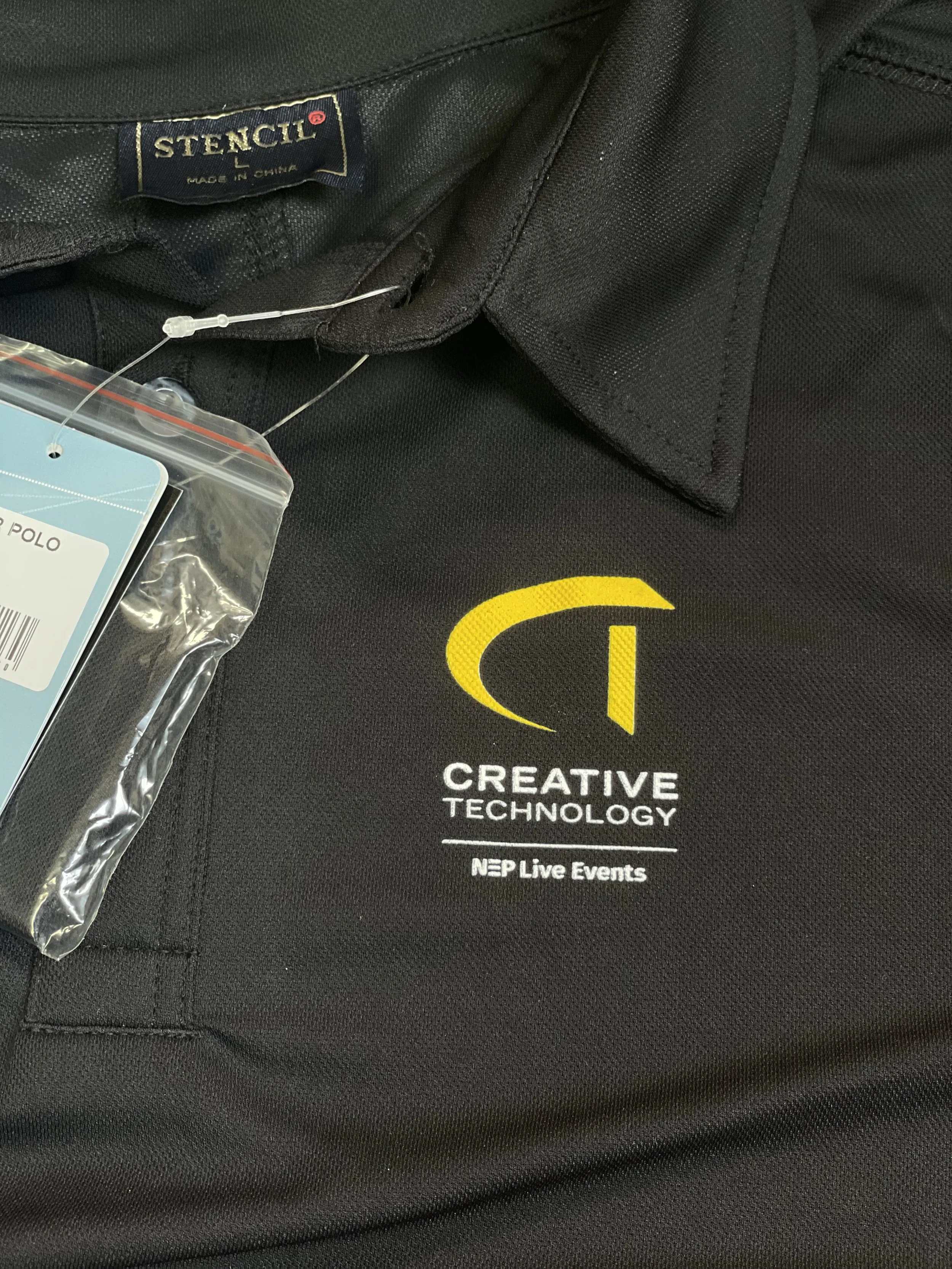 Close-up of a black polo shirt with a yellow and white logo and text that reads 'CREATIVE TECHNOLOGY NPLive Events', with a tag that says 'STENCIL' and 'Made in China'.