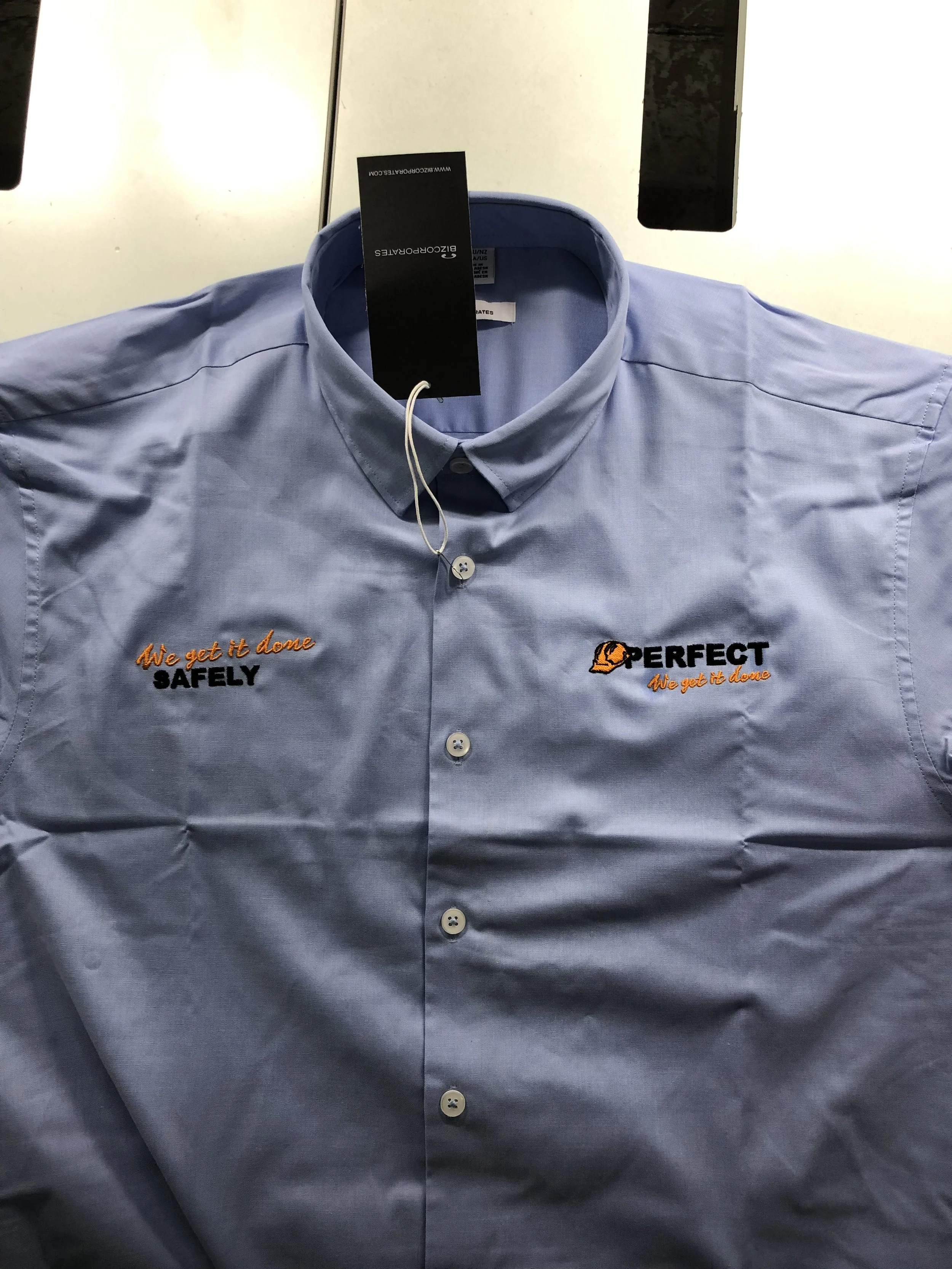 A light blue work shirt with embroidered text and logo, featuring the phrase 'We get it done SAFELY' on the left chest and the phrase 'PERFECT We get it done' with an orange helmet logo on the right chest, laid flat on a surface.