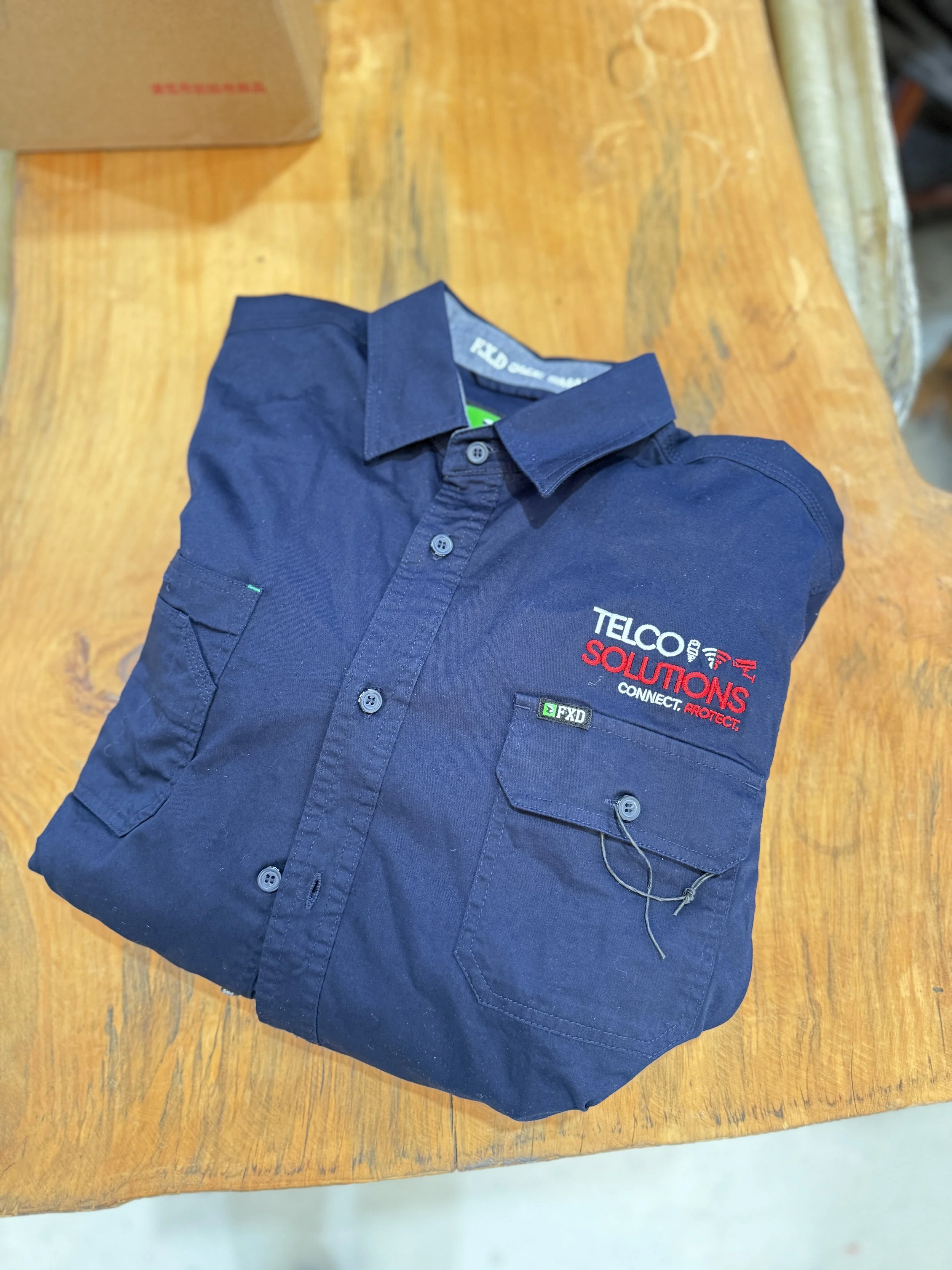 Folded navy blue work shirt with embroidery that says "TELCO SOLUTIONS CONNECT. PROTECT." and a small pocket on the left chest with a wire attached.