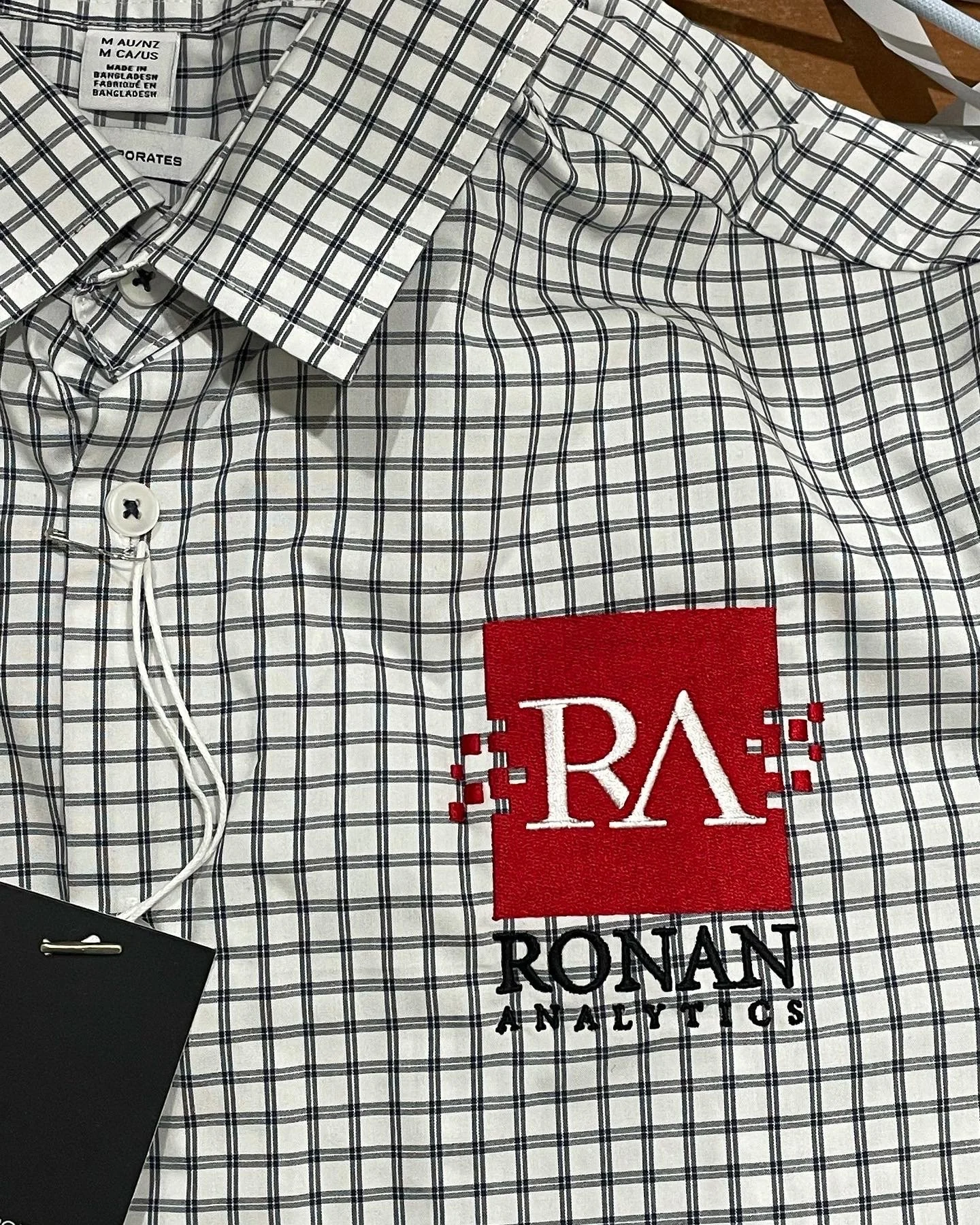 A white dress shirt with a black checkered pattern and a red embroidered patch that reads 'RA' with the words 'RONAN ANALYTICS' below it, displayed on a flat surface.