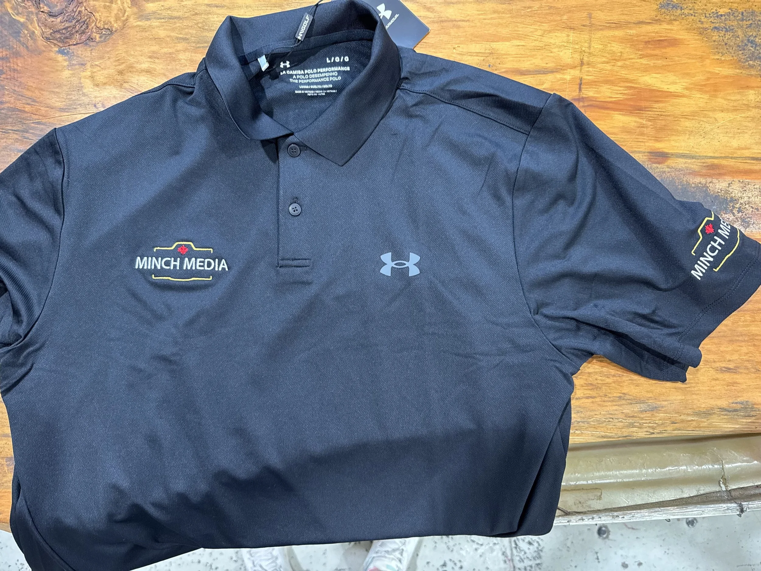 Black Under Armour polo shirt with embroidered 'Minch Media' logo on the left chest and right sleeve, laid flat on a wooden surface.
