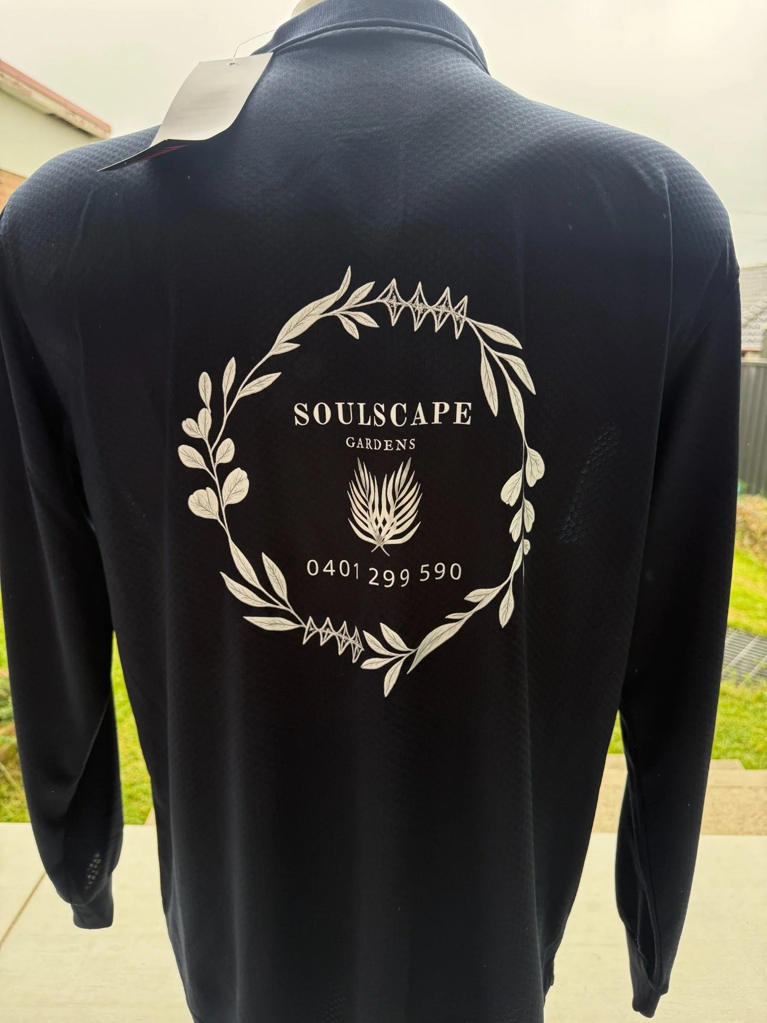 Black sports shirt with white logo and text for 'Soulscape Gardens,' featuring a wreath of leaves, a central plant or flower, and a phone number.
