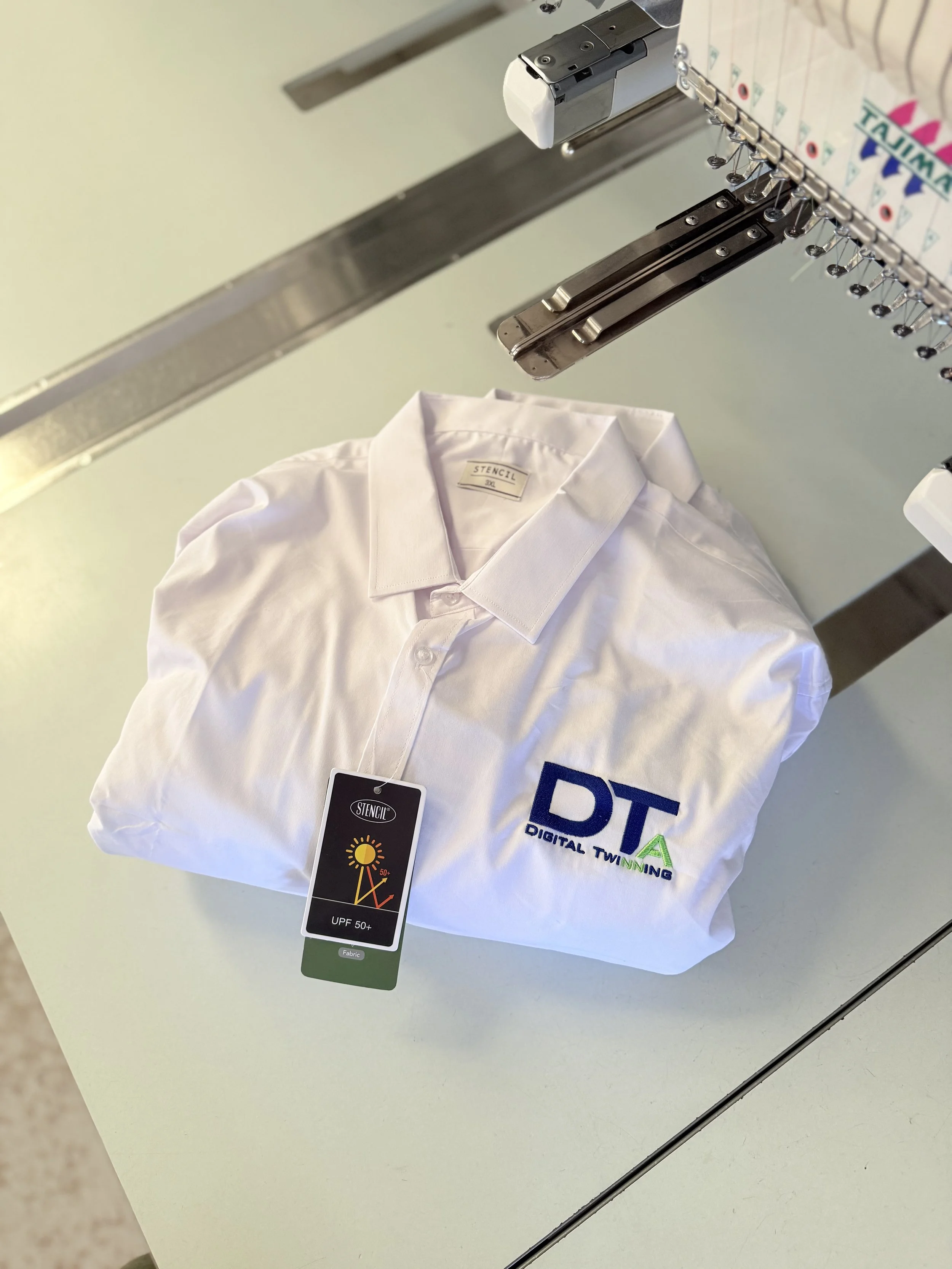 White polo shirt with embroidered logo and a tag, placed on a white surface, with embroidery machines visible above.