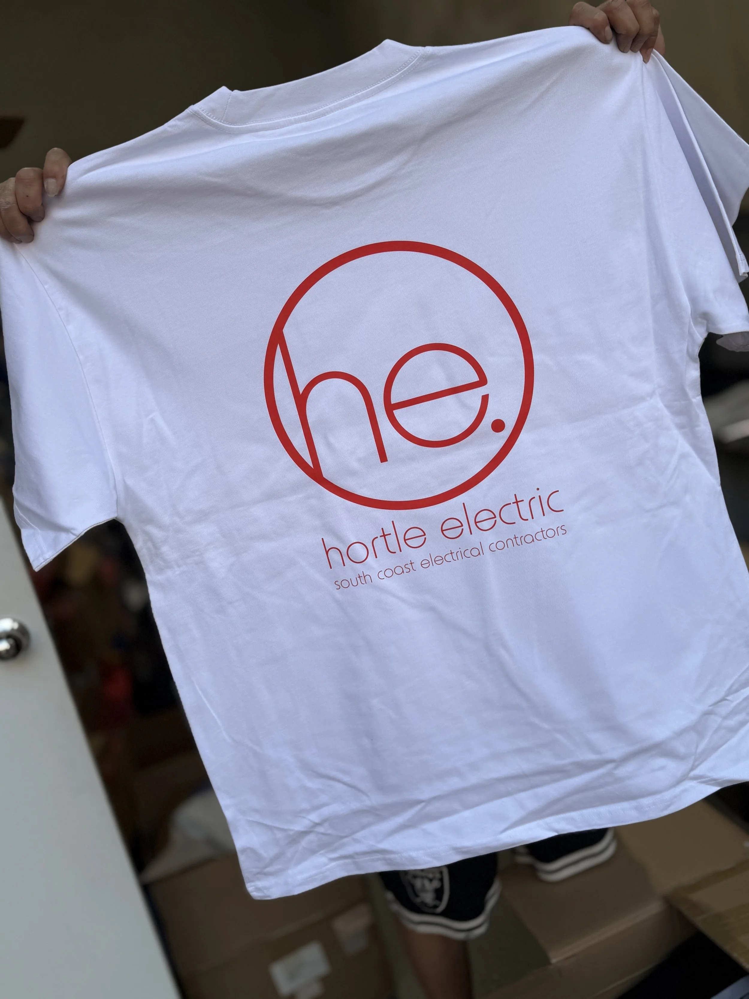 White T-shirt with a red circle design and the word "he" inside in red outline. Below the circle, it says "hortle electric" in red text, with smaller text underneath reading "south coast electrical contractors." Someone is holding the shirt up by the