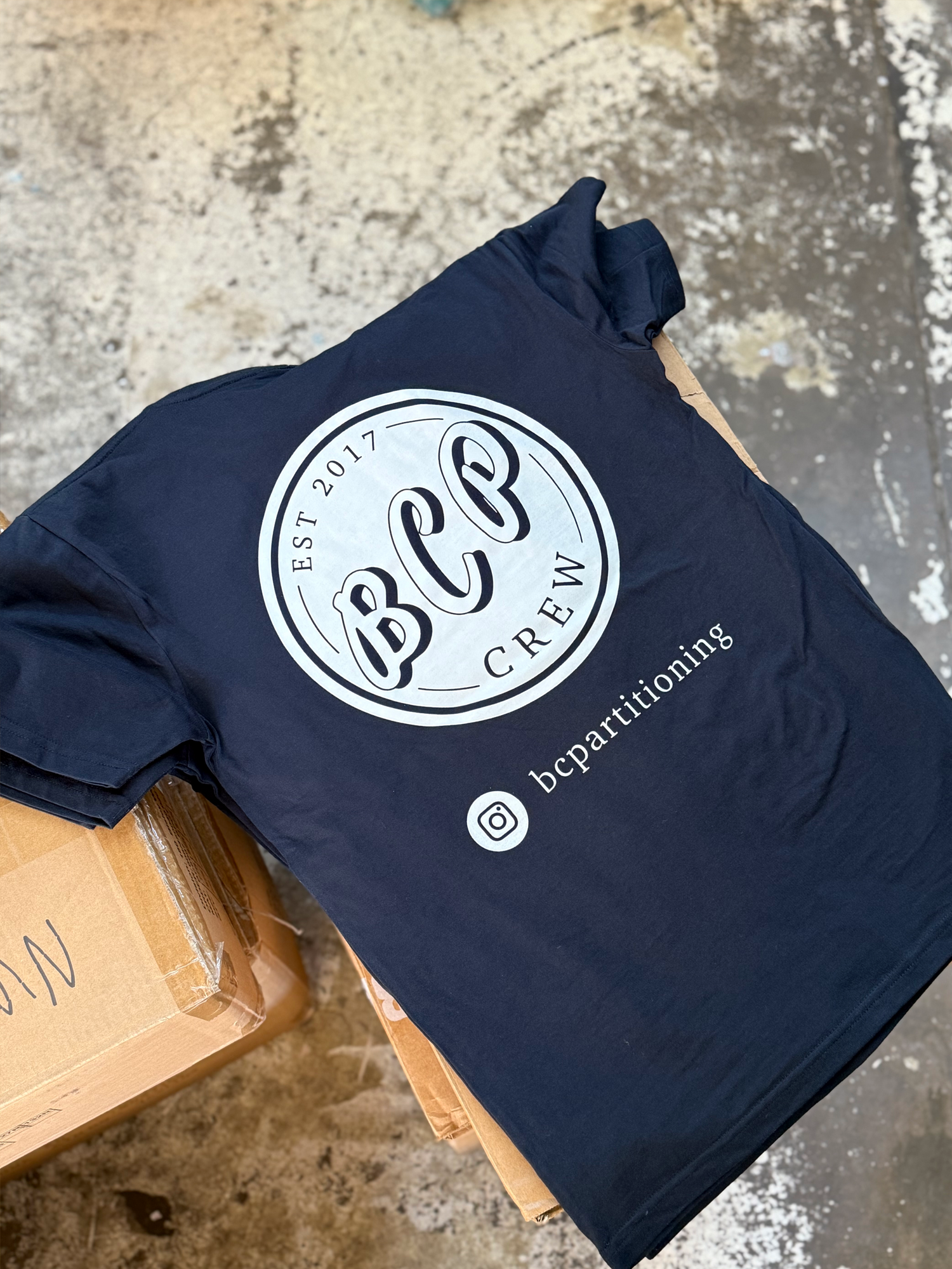 A black screen printed t-shirt with the logo of BCP Crew, established in 2017, printed in white on the front, laying on a cardboard box.