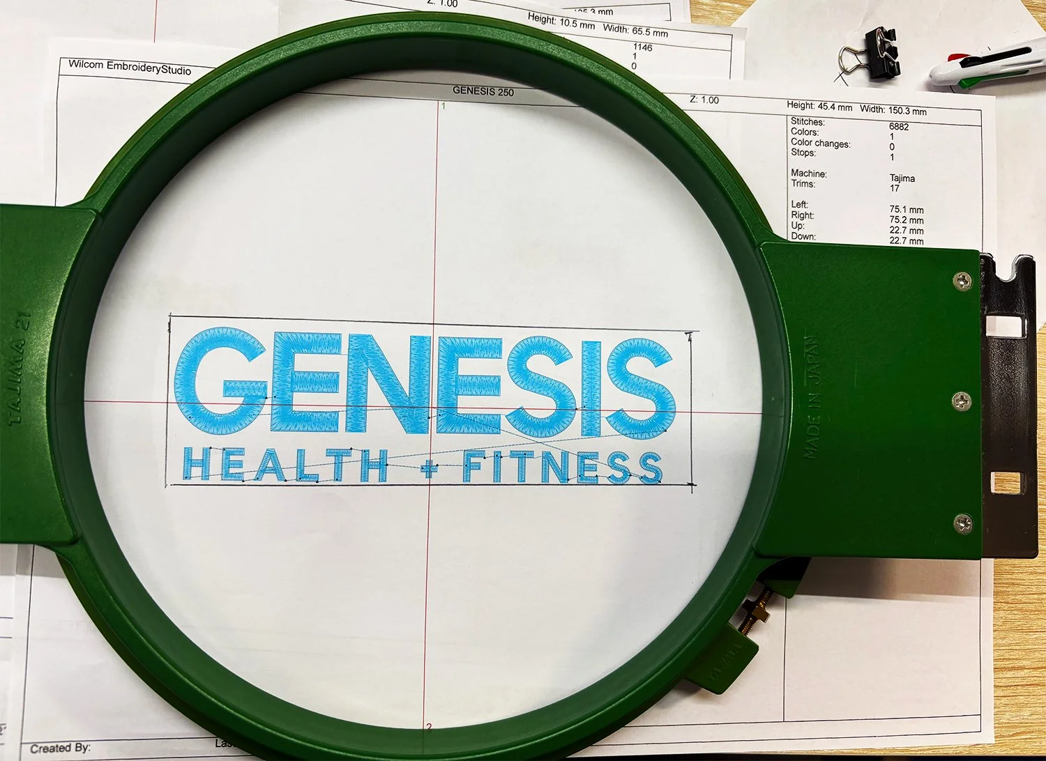 Embroidery hoop with blue embroidered text reading 'GENESIS HEALTH + FITNESS' placed over design and sewing patterns and papers.